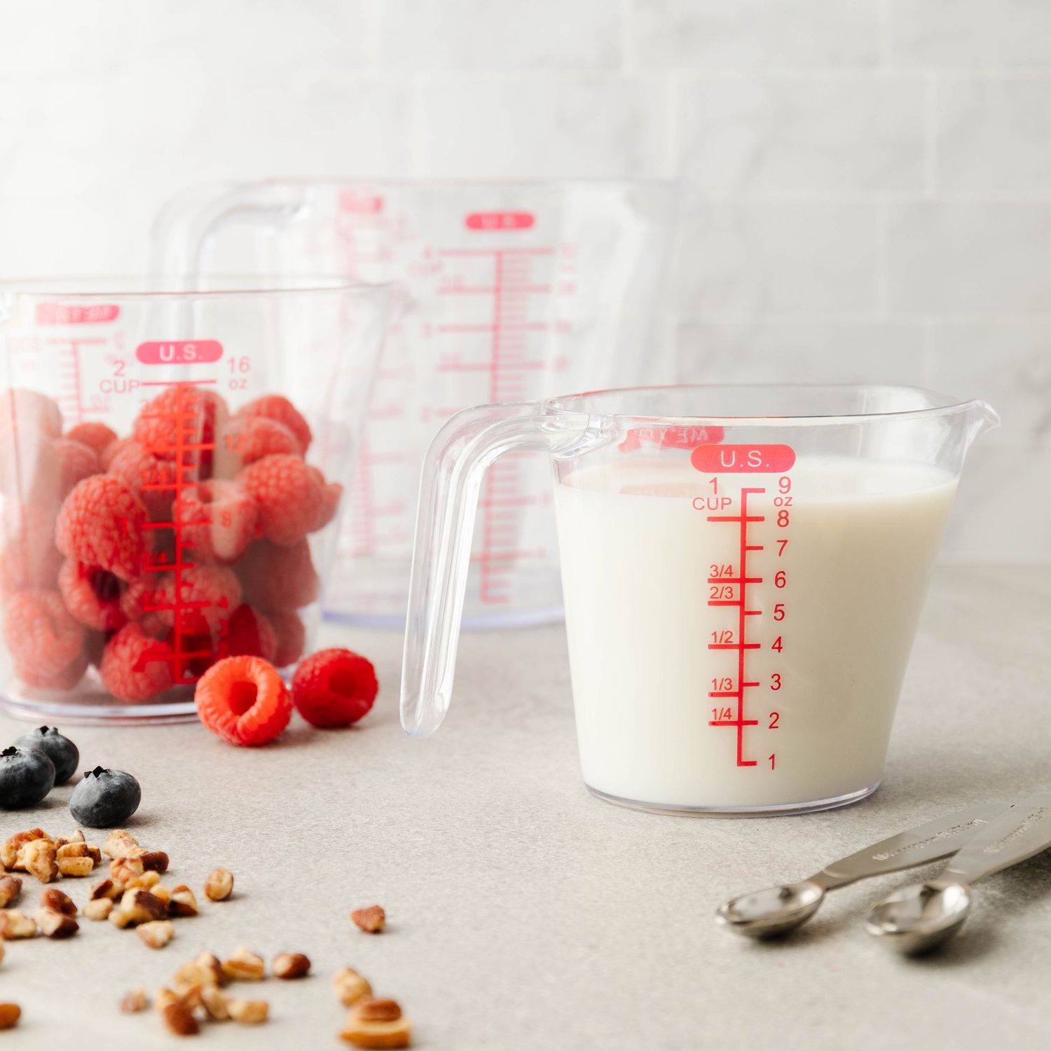 Two clear measuring cups filled with raspberries and milk alongside blueberries and granola on a light background.