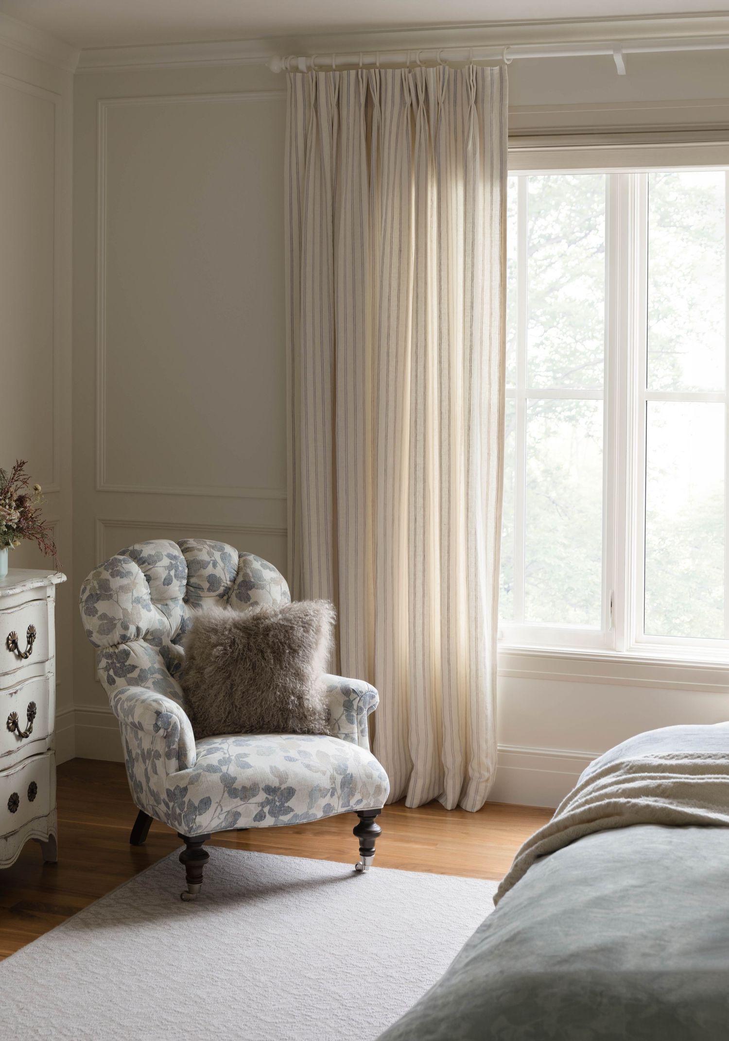 Elegant bedroom corner with tufted floral armchair, fur pillow, sheer curtains, and French provincial dresser.