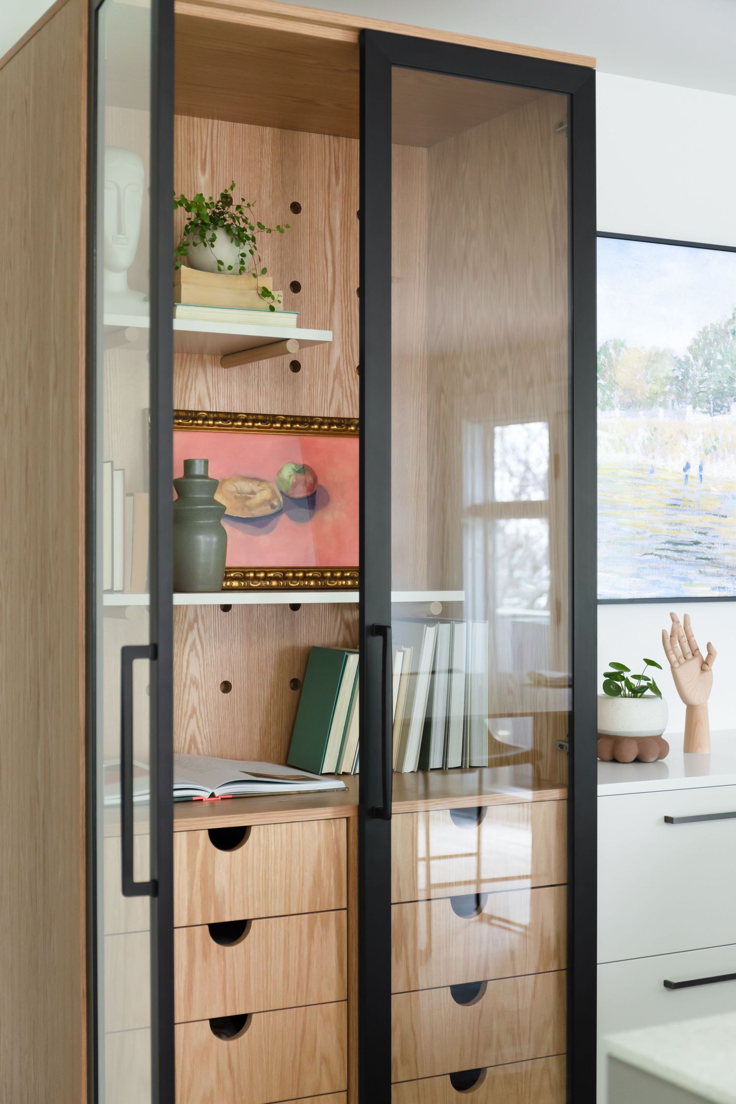 Contemporary built-in cabinet with black metal frame, glass doors, light wood shelving displaying books, plants, and artwork.