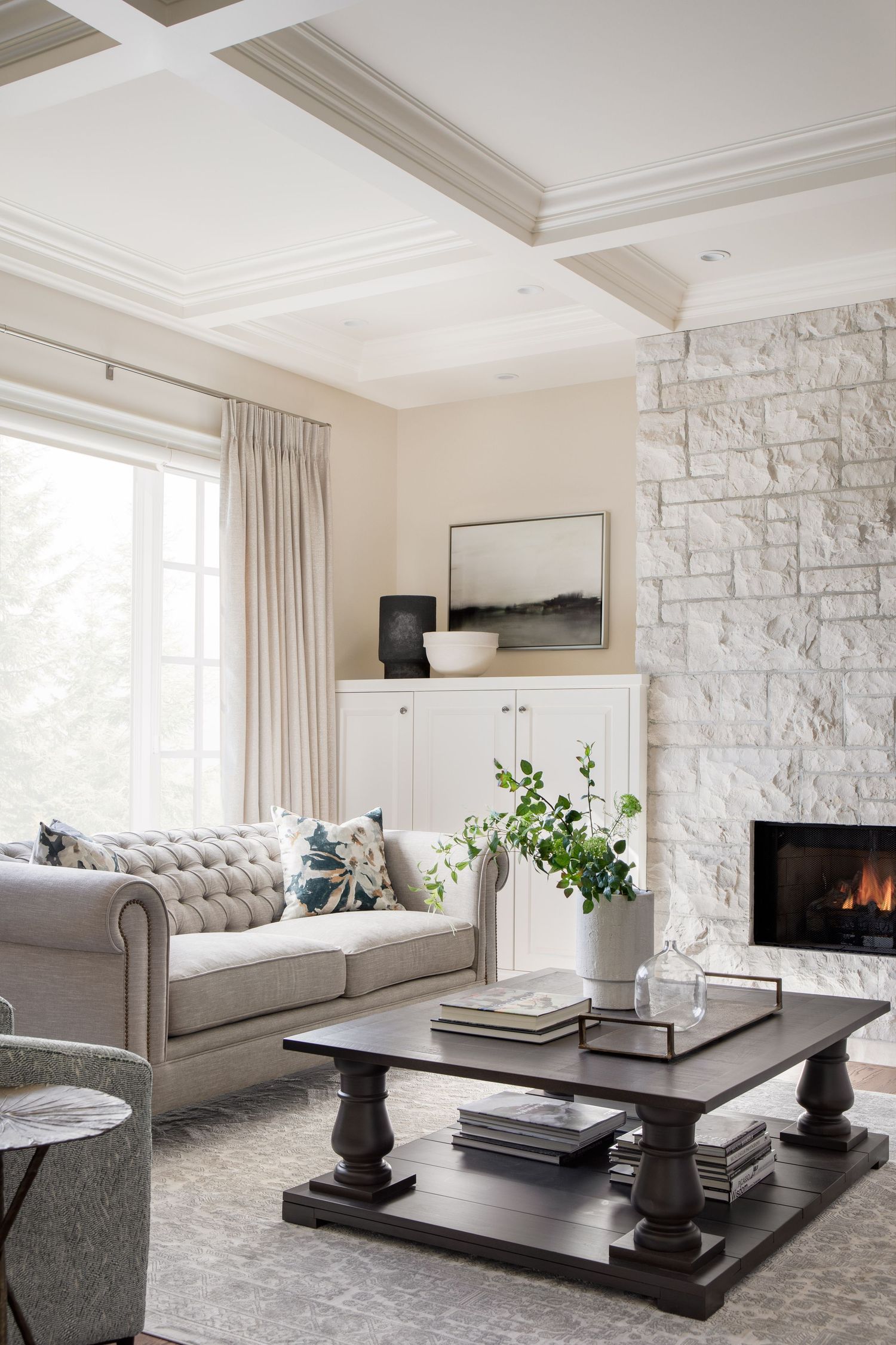 Bright living room with tufted sofa, stone fireplace, coffered ceiling, and dark wood coffee table.