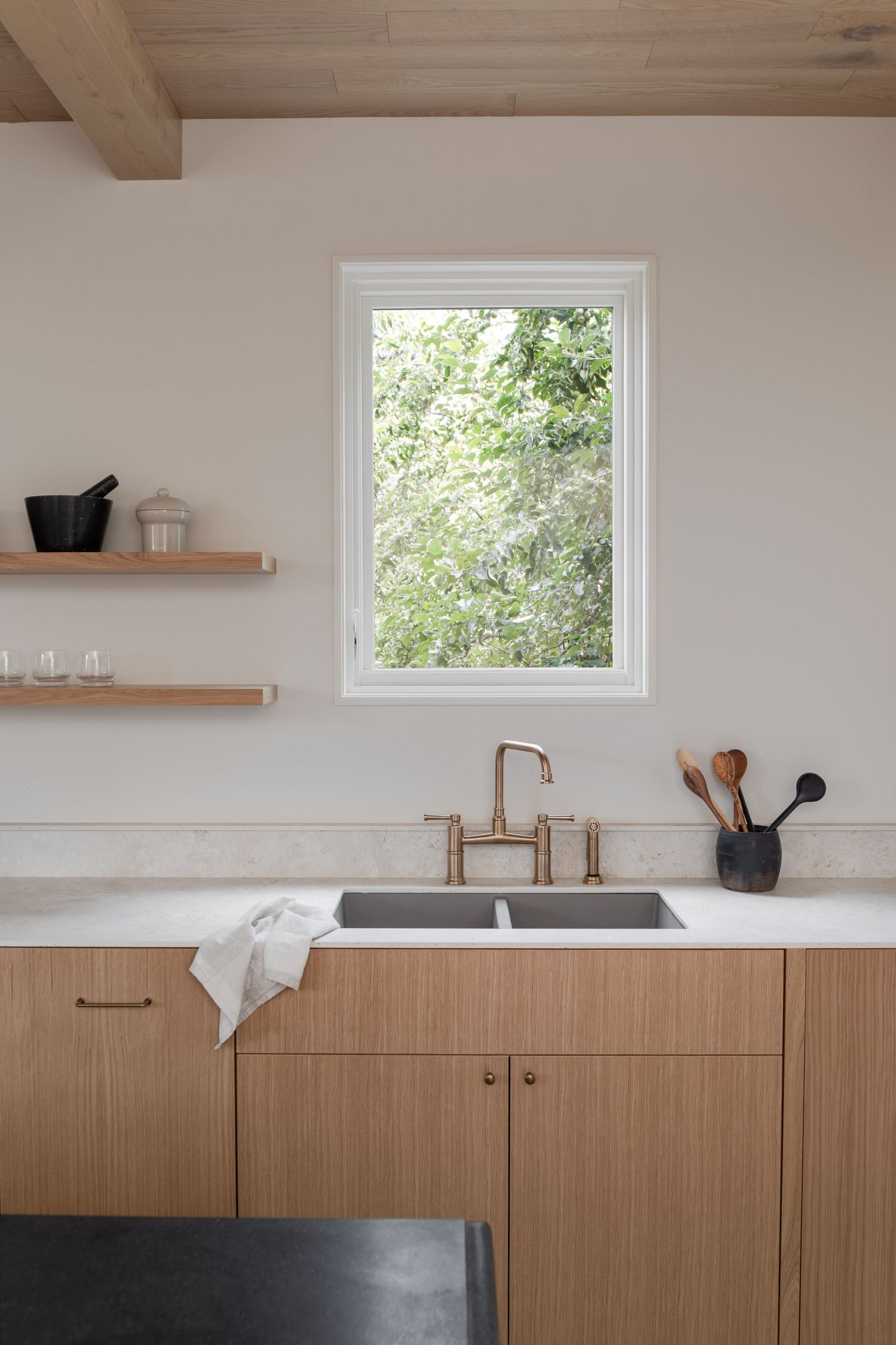 Minimalist oak kitchen with double sink, rose gold bridge faucet, open shelving, and garden window view.