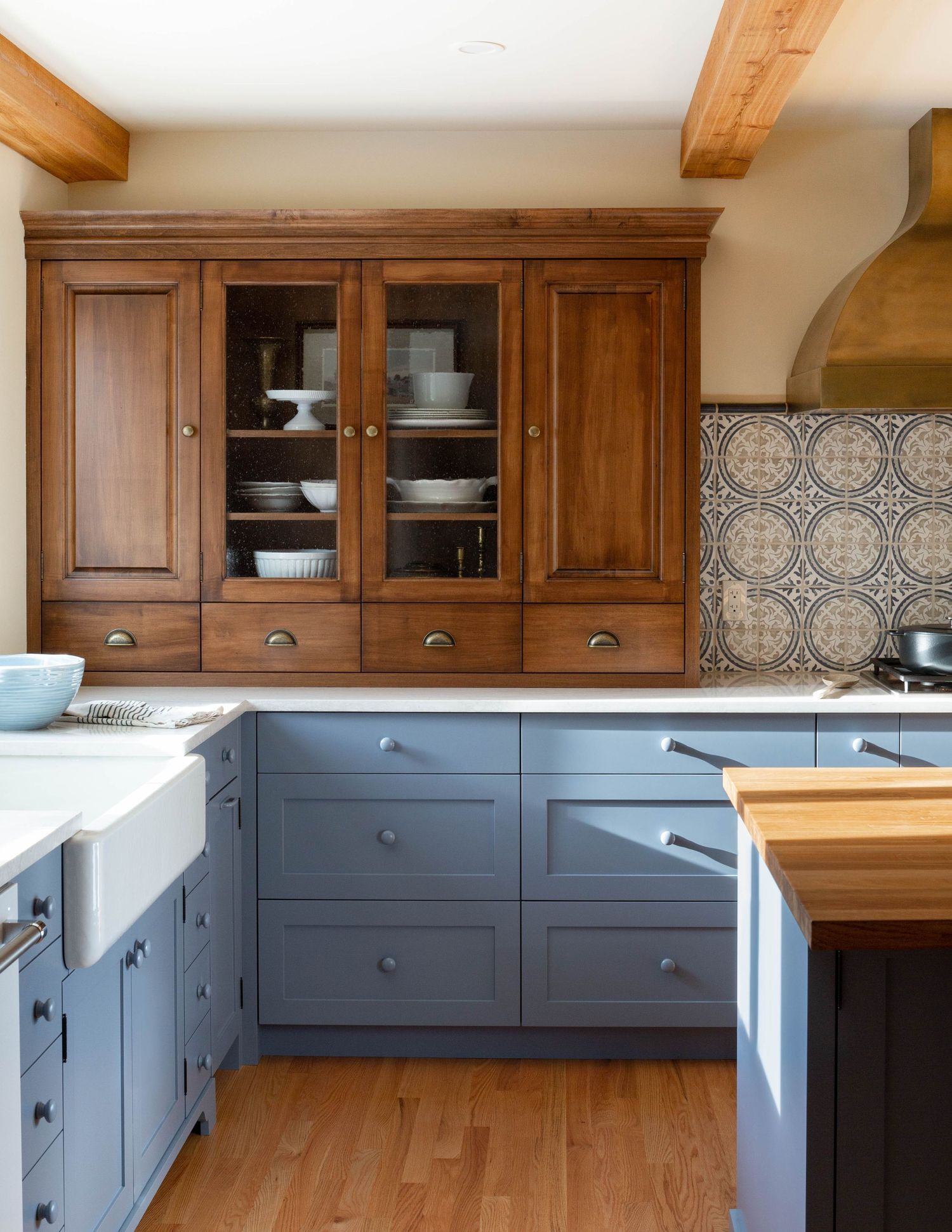 Farmhouse kitchen featuring blue-gray lower cabinets, natural wood upper cabinets, patterned tile backsplash, and exposed ceiling beams.