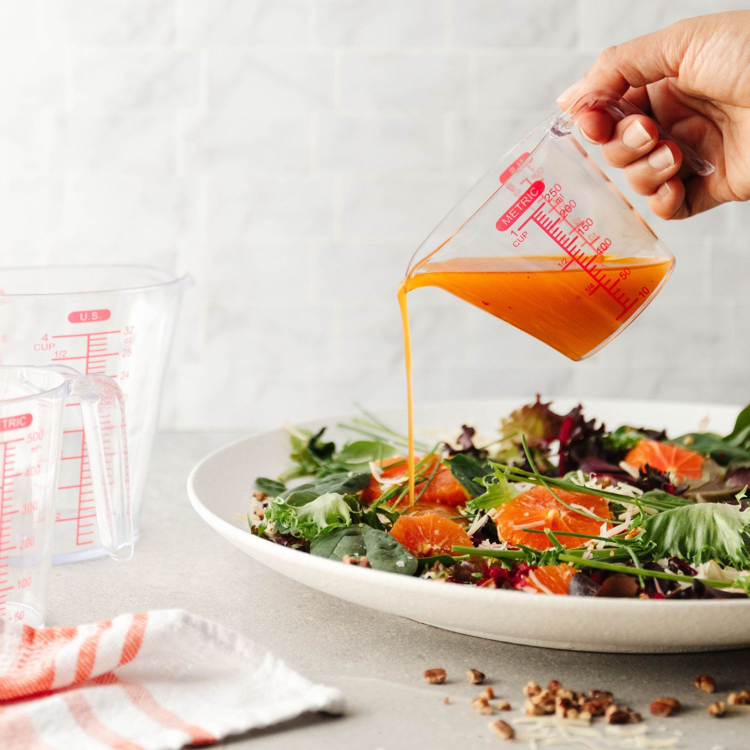 Hand pouring orange vinaigrette dressing from a measuring cup onto a fresh mixed green salad.