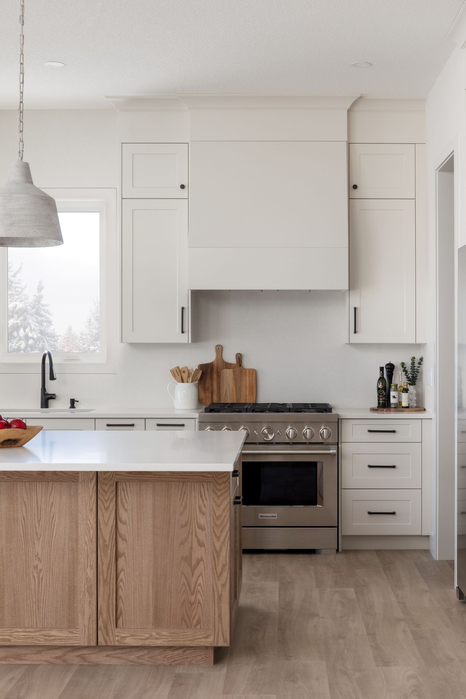 White shaker kitchen with wood island, custom hood, stainless range, and concrete pendant light.