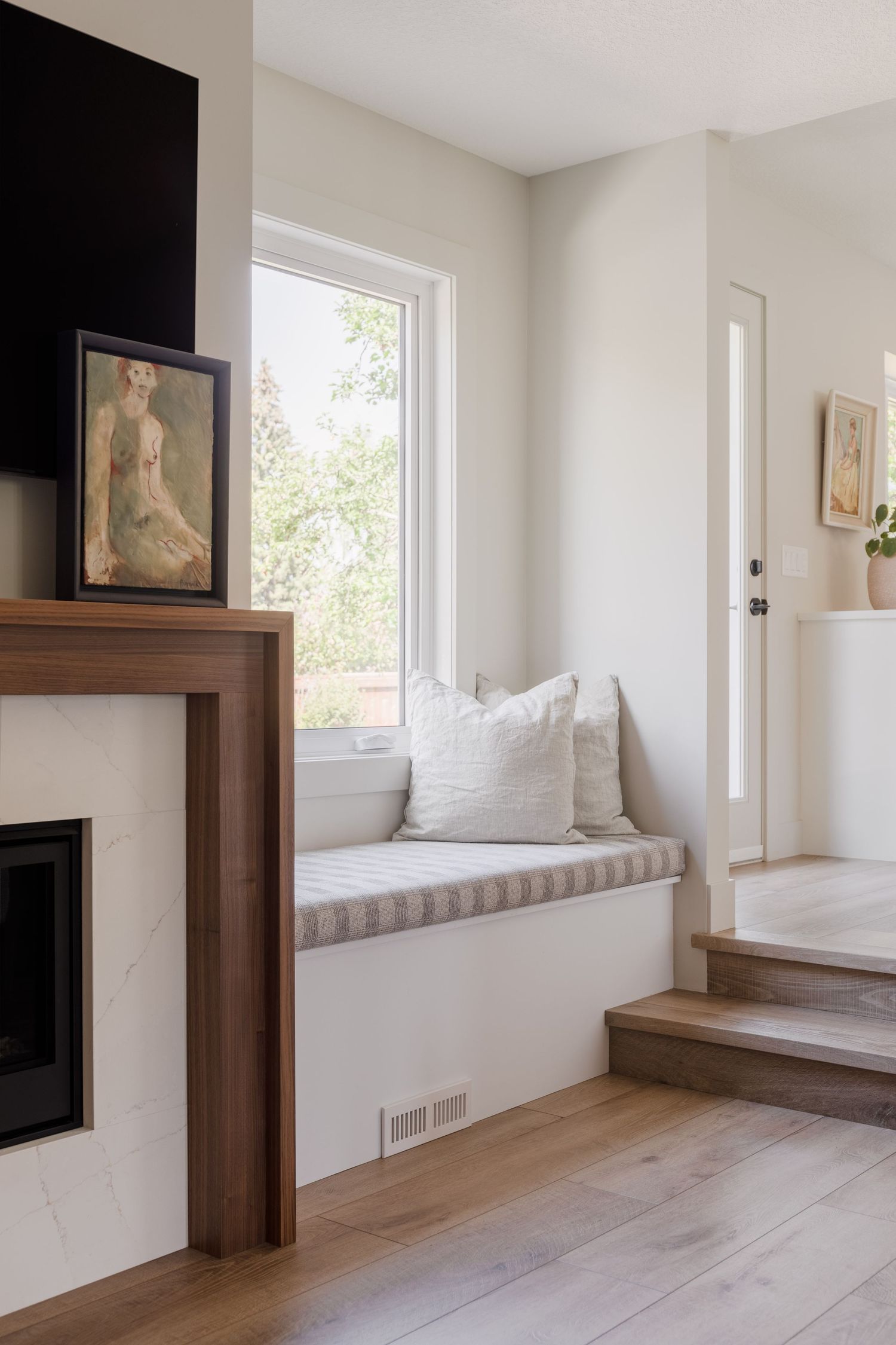 Minimalist living space with white window seat, linen pillows, light wood floors, walnut fireplace surround, and white marble hearth.