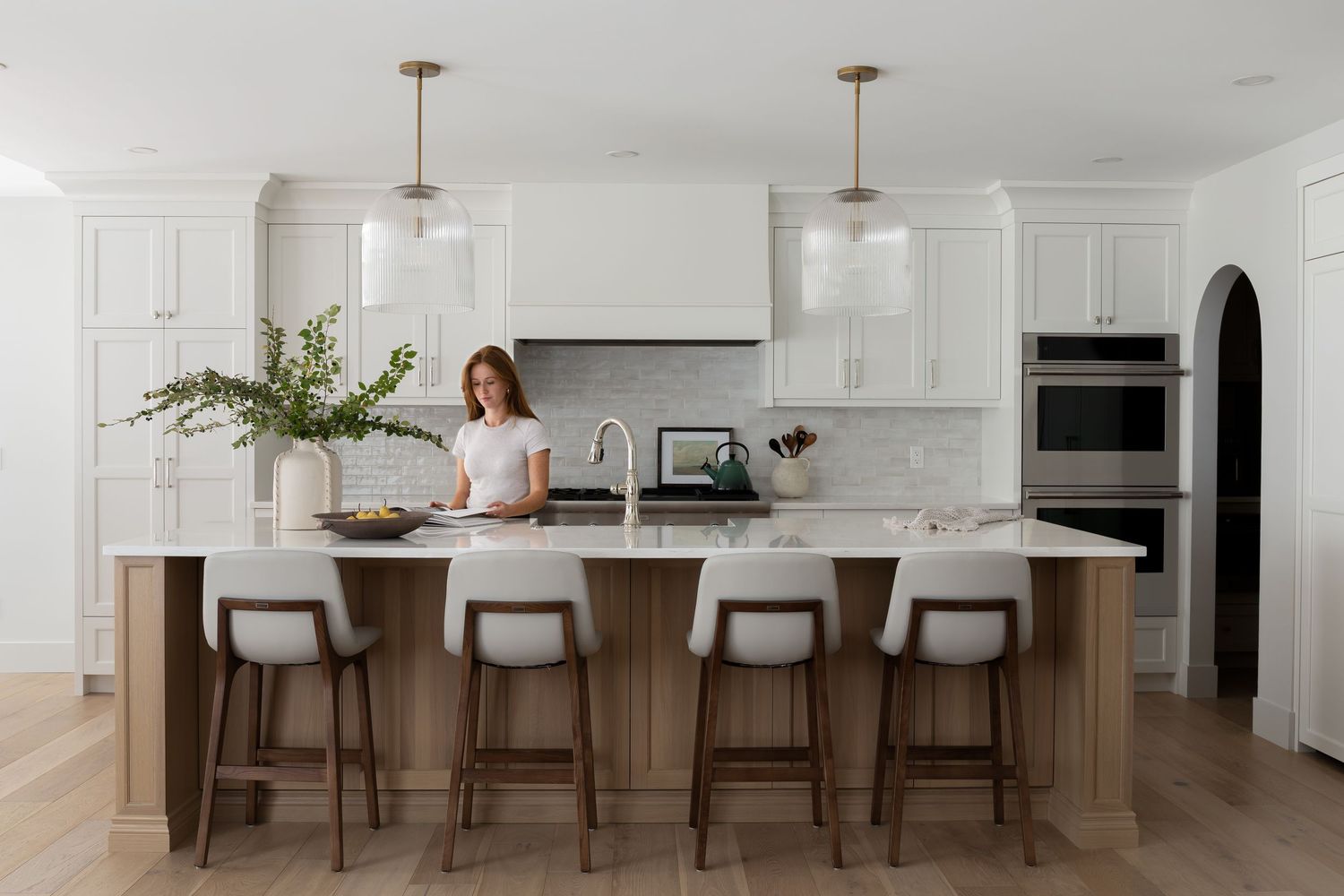 Elegant white shaker kitchen with large wood island, white countertops, glass pendants, and arched doorway detail.