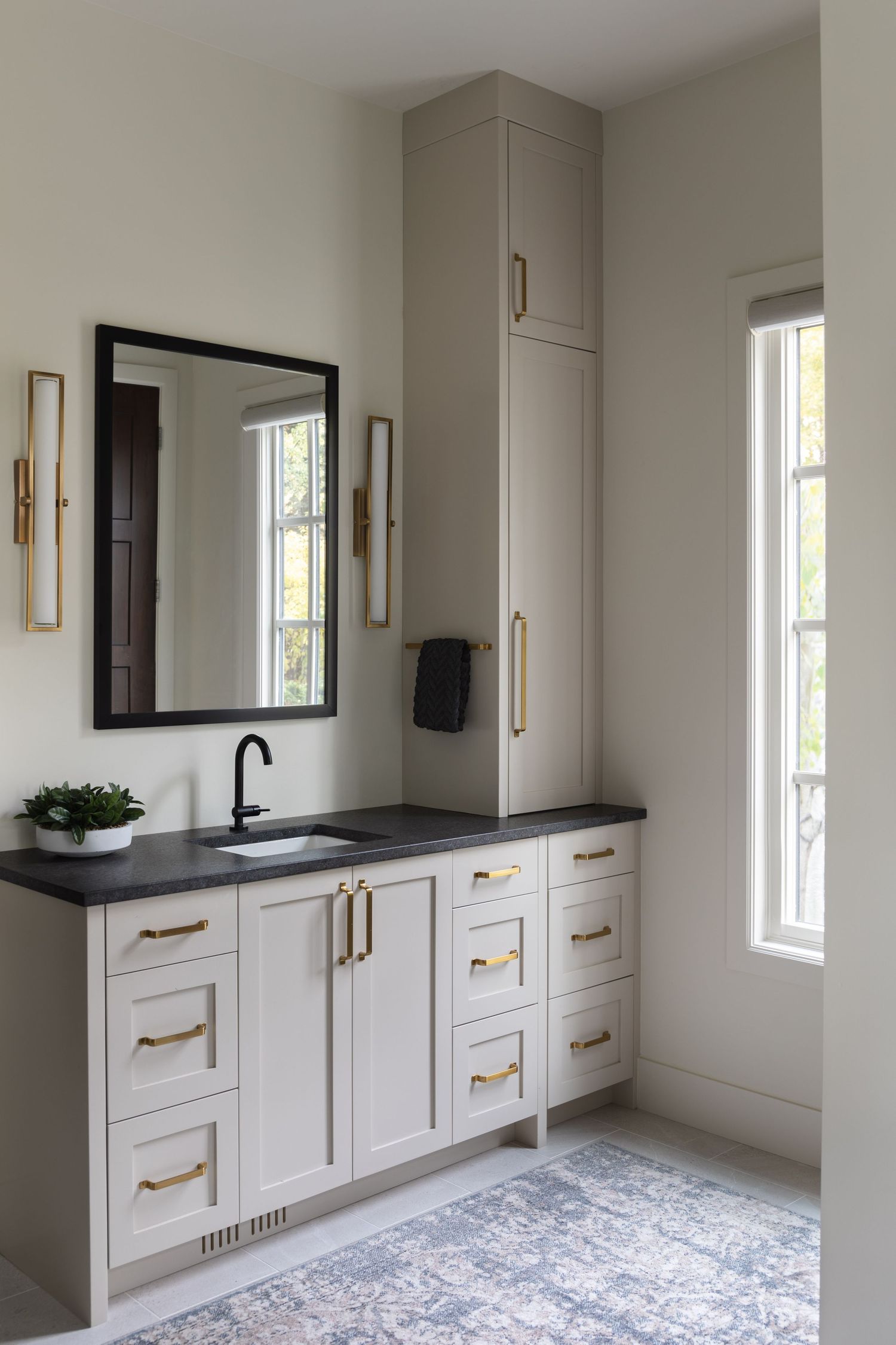 Transitional bathroom vanity with gray cabinets, brass hardware, black countertop, tall linen tower, and patterned area rug.