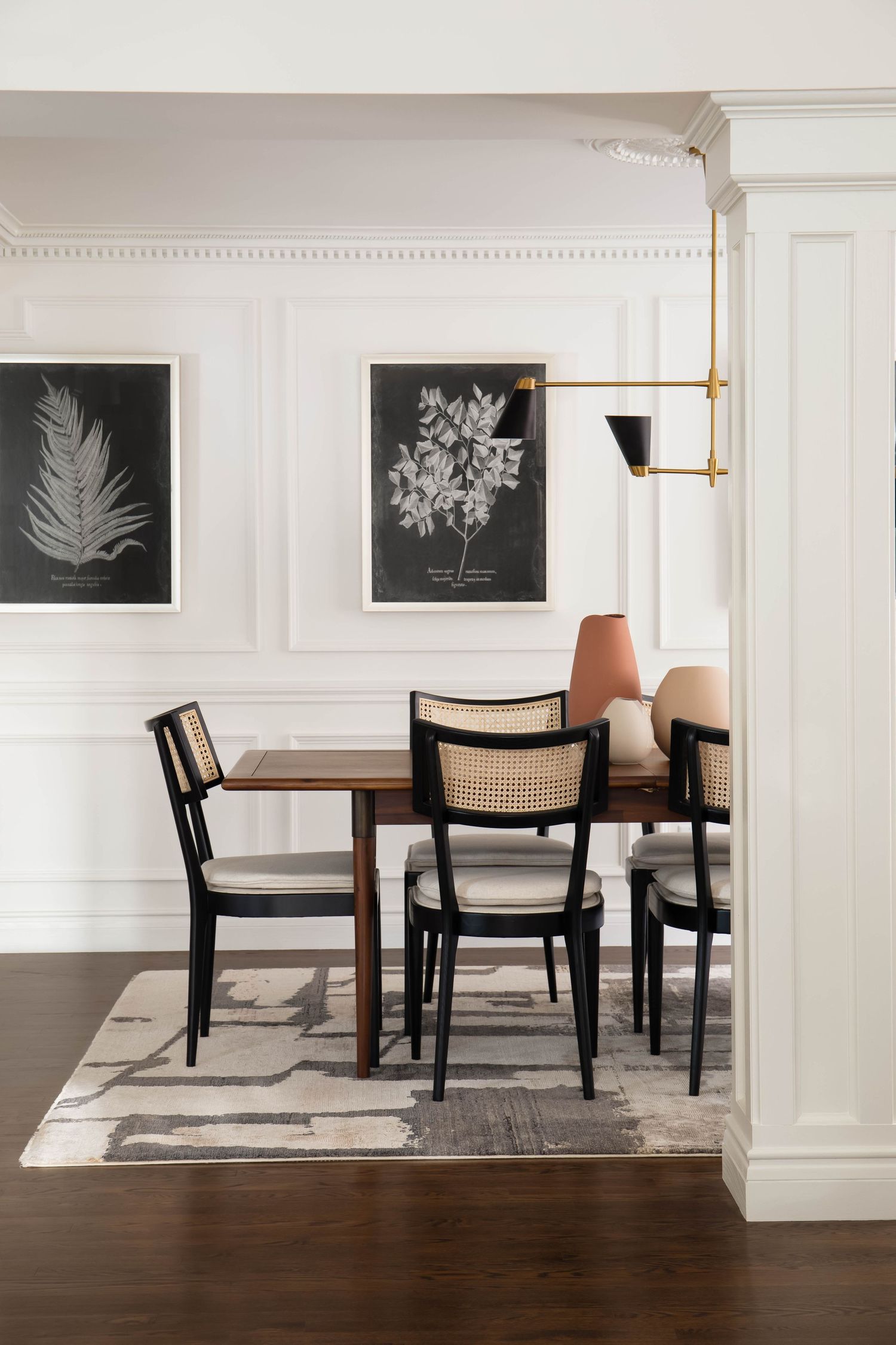 Dining nook with black cane chairs, wood table, botanical prints, white paneling, and brass swing-arm pendant.