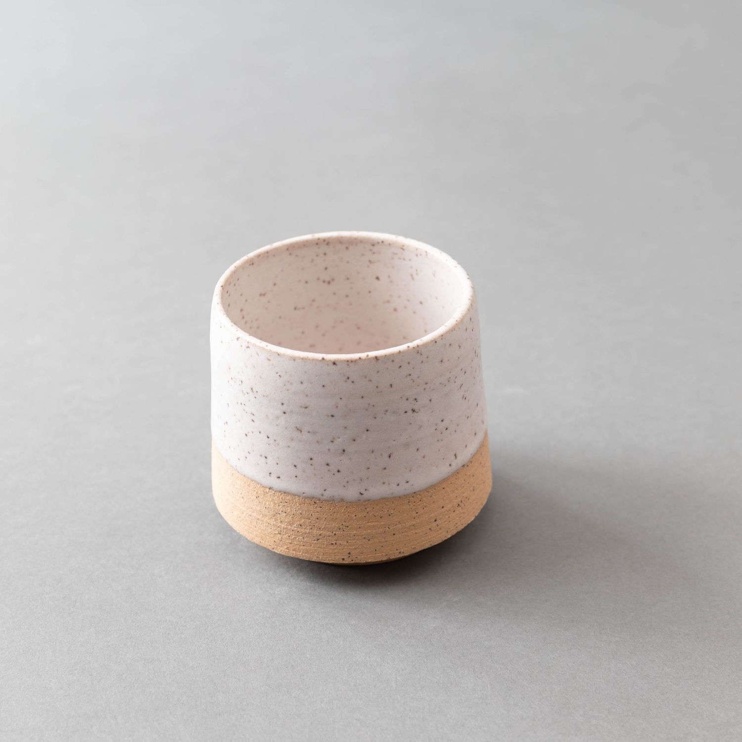 Single minimalist two-tone ceramic cup with white speckled glaze and natural unglazed clay base.