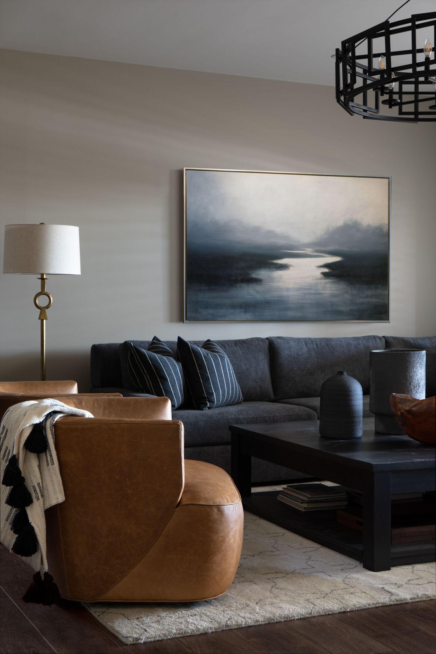 Moody living room with dark sectional sofa, leather swivel chair, abstract landscape art, and black chandelier.