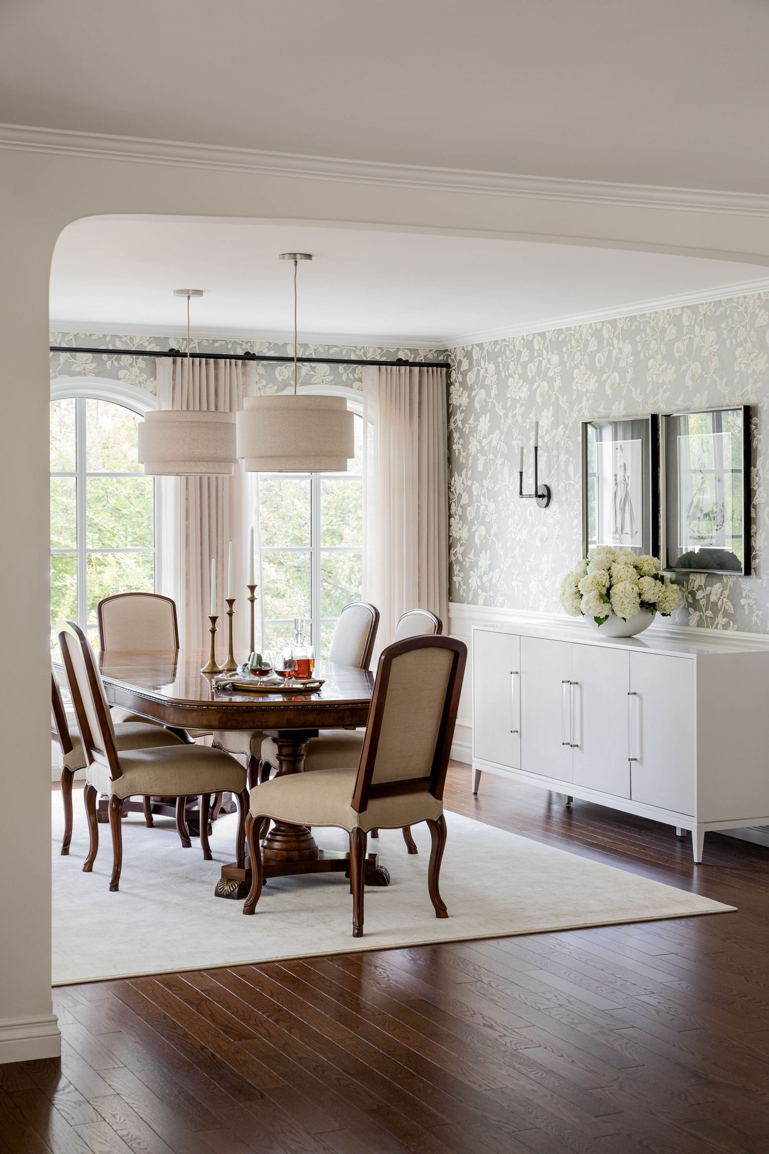 Elegant dining room with round wood table, upholstered chairs, floral wallpaper, and twin drum pendant lights.