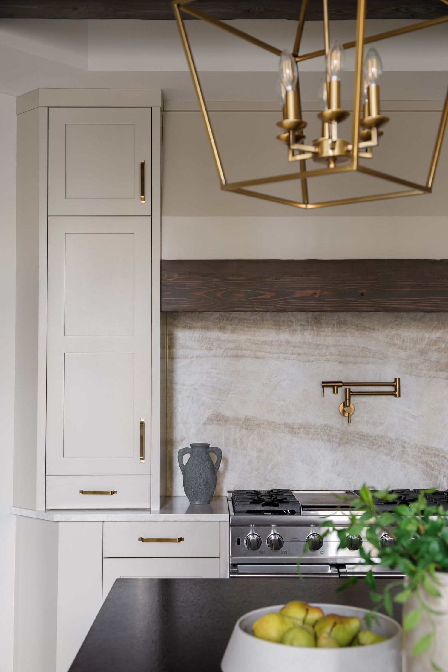 Kitchen detail showing cream shaker cabinets, brass hardware, stone slab range backsplash, pot filler, and gold geometric pendant.