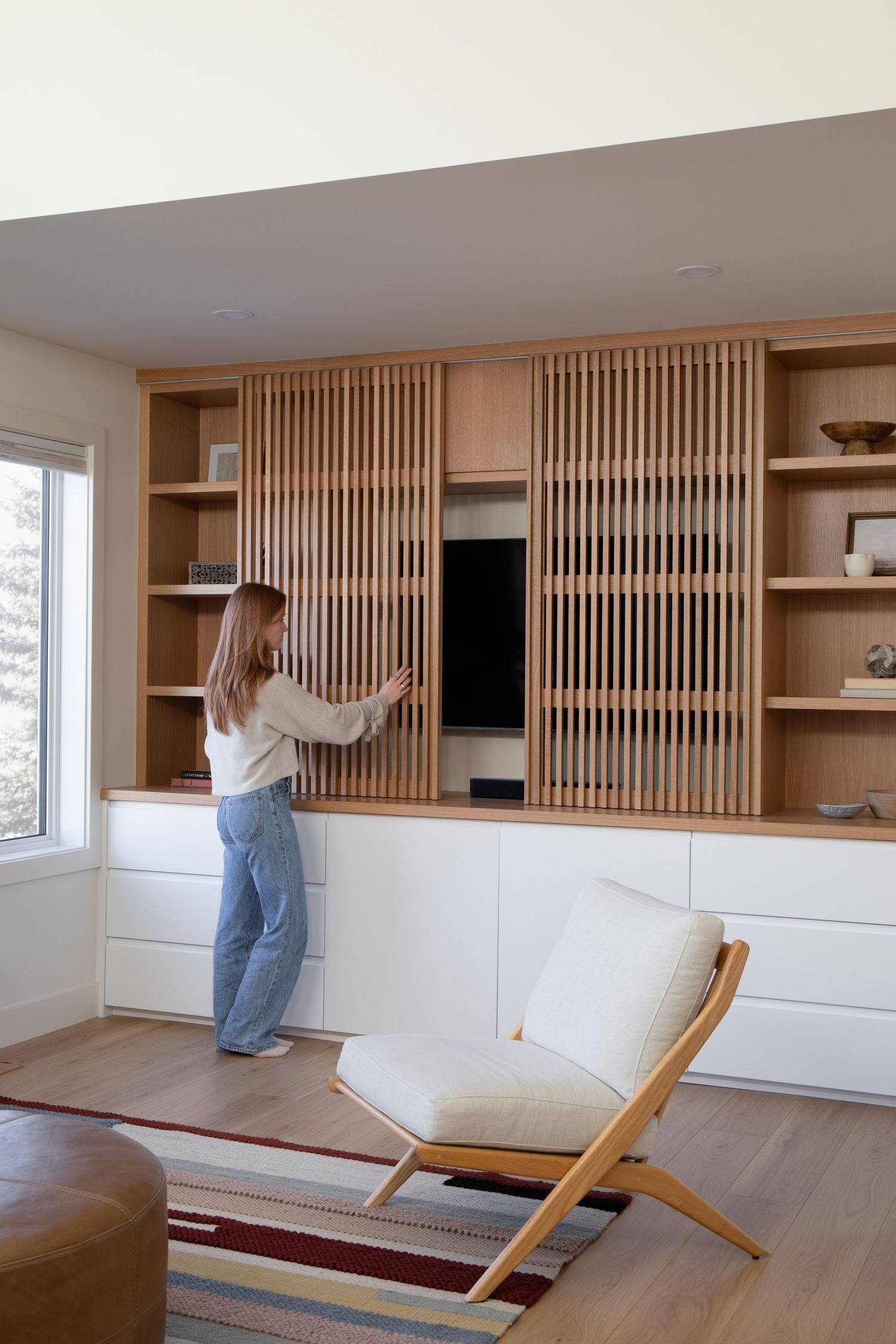 Woman opening slatted oak wood built-in shelving unit concealing TV in a bright modern living room.
