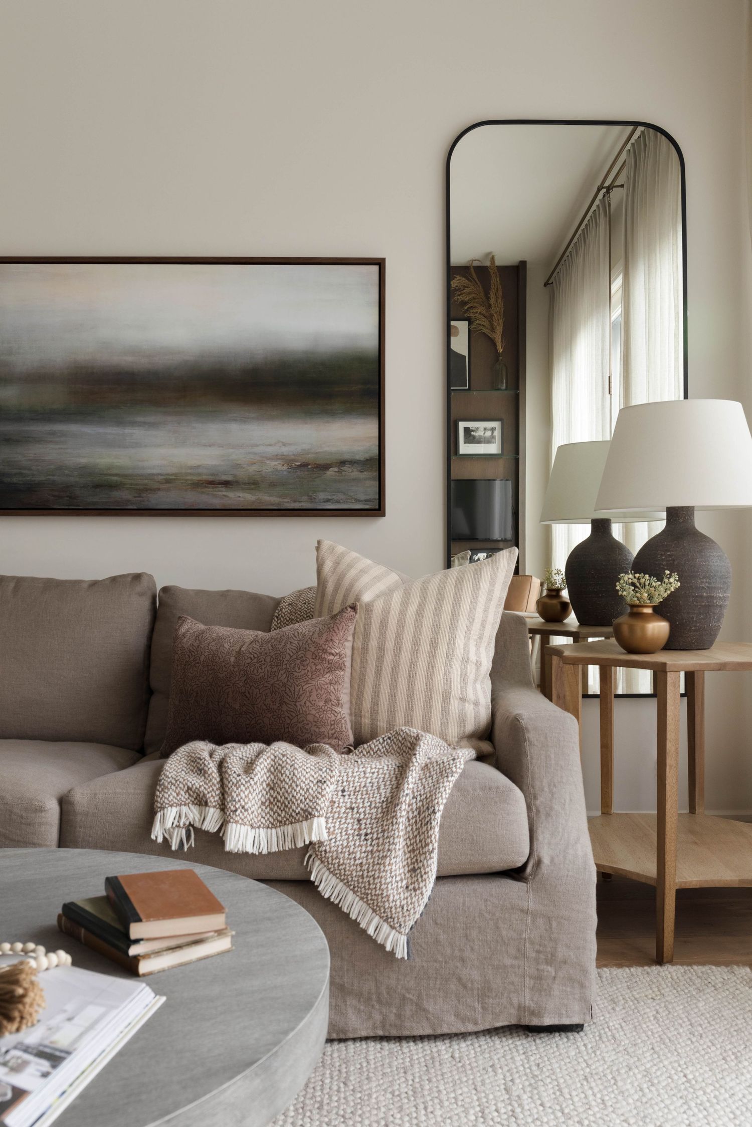 Neutral living room with linen sofa, layered pillows, arched mirror, and abstract wall art.