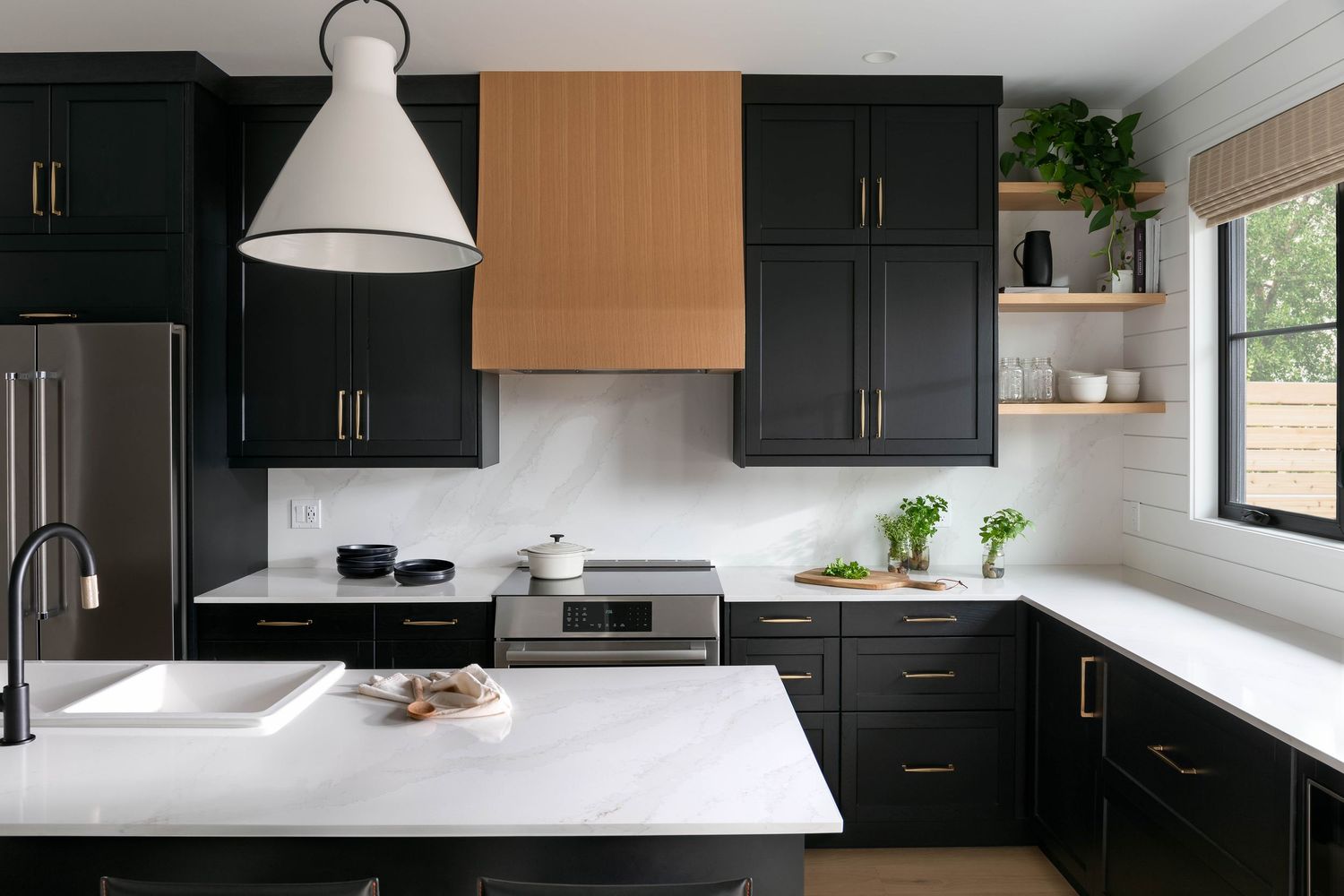 Modern kitchen with matte black cabinets, wood range hood, white marble countertops, white pendant light, and stainless steel appliances.