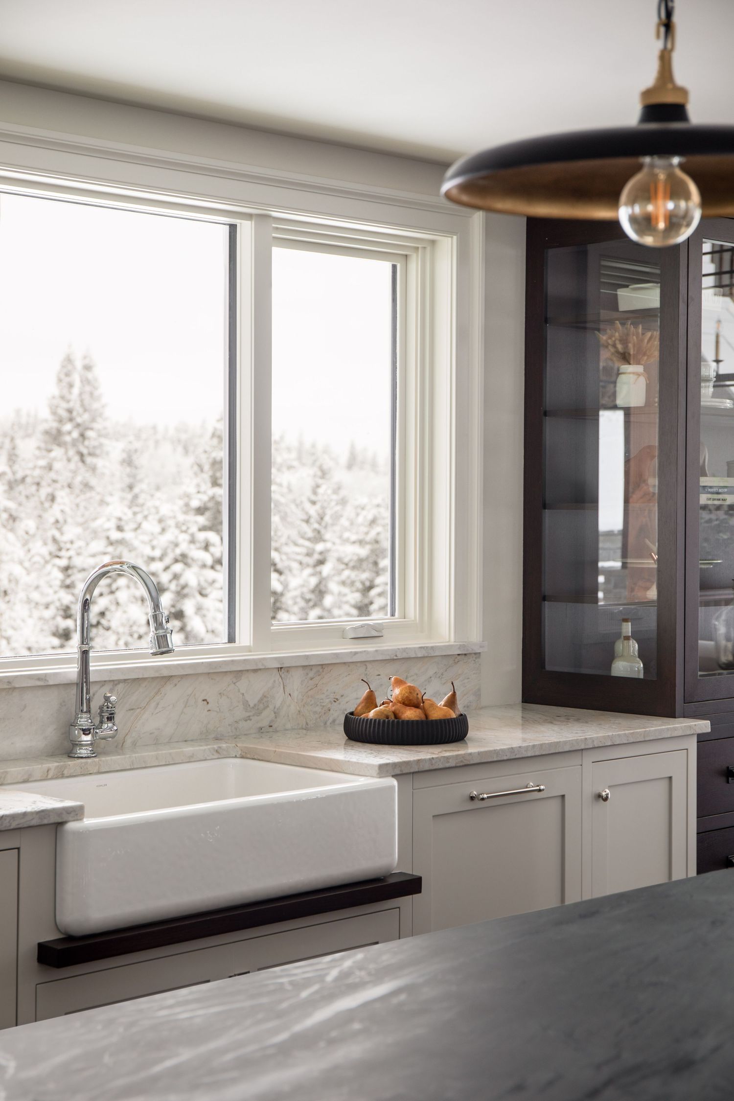 Farmhouse kitchen sink with marble countertop, gray cabinets, chrome faucet, and snowy winter view through large window.