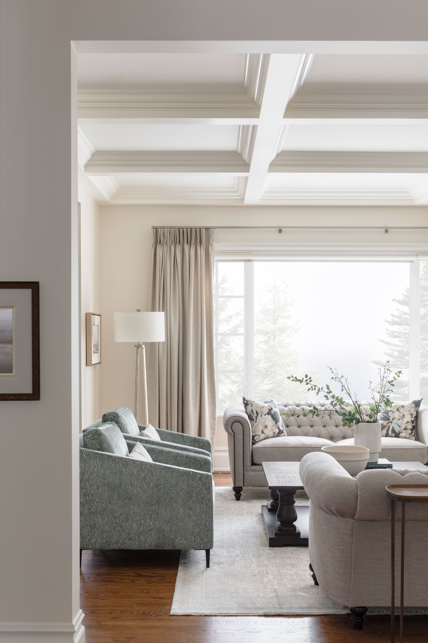 Elegant living room with coffered ceiling, tufted sofa, sage green armchairs, and large window with linen drapes.