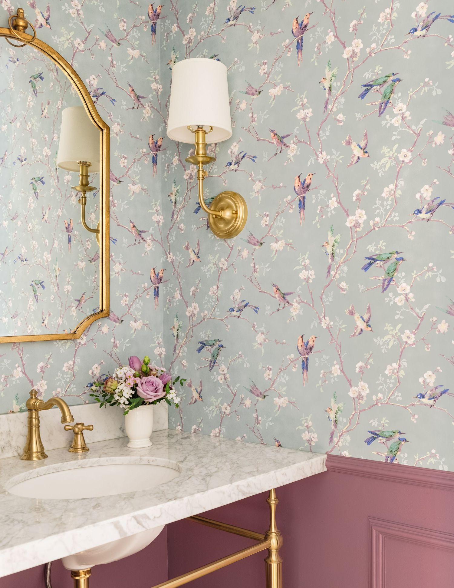 Powder room with chinoiserie bird wallpaper, brass fixtures, arched gold mirror, and pink wainscoting.