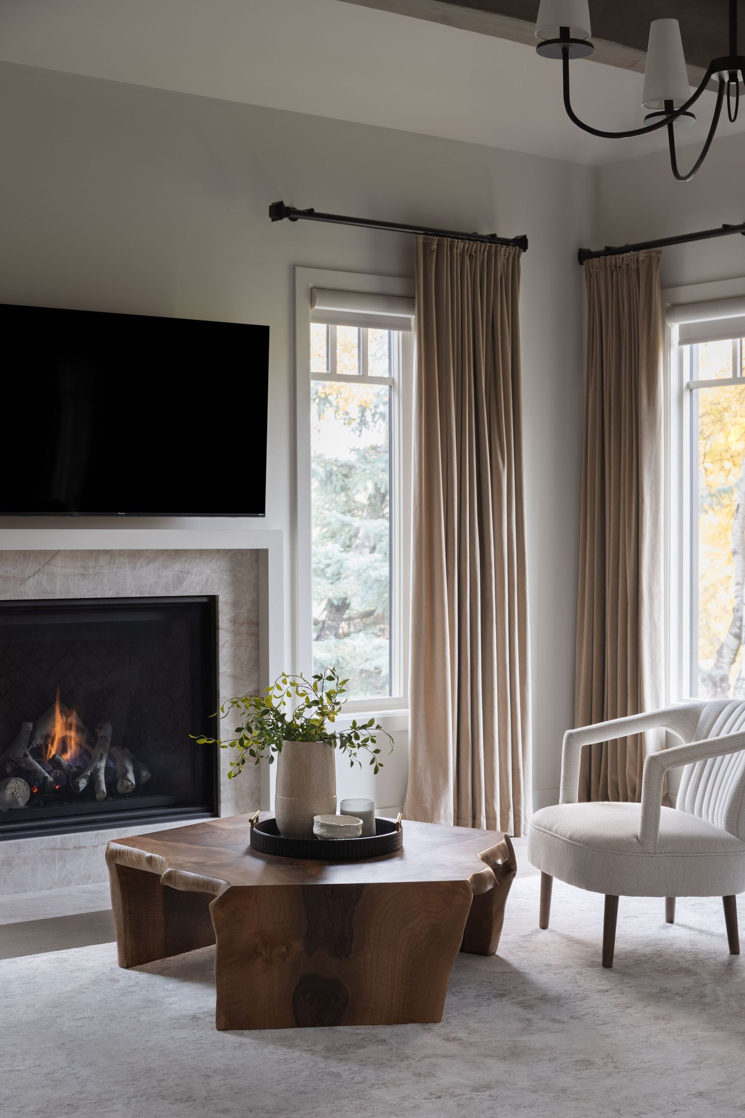 Cozy living room with live-edge wood coffee table, marble fireplace, wall-mounted TV, linen drapes, and neutral upholstered chair.