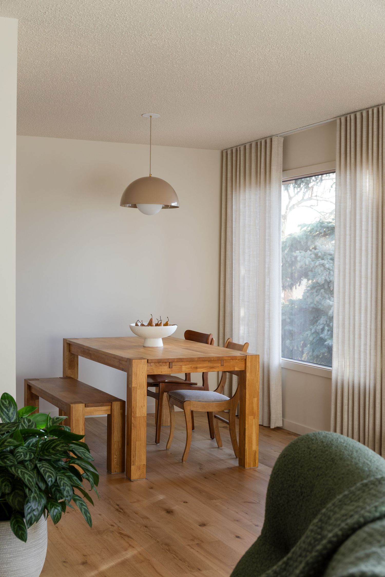 Simple dining area with solid wood table, bench, dome pendant, linen curtains, and green potted plant.