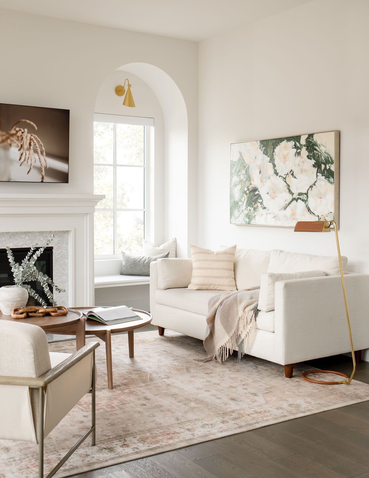 Airy living room featuring white sofa, floral art, arched window, fireplace, and blush vintage rug.