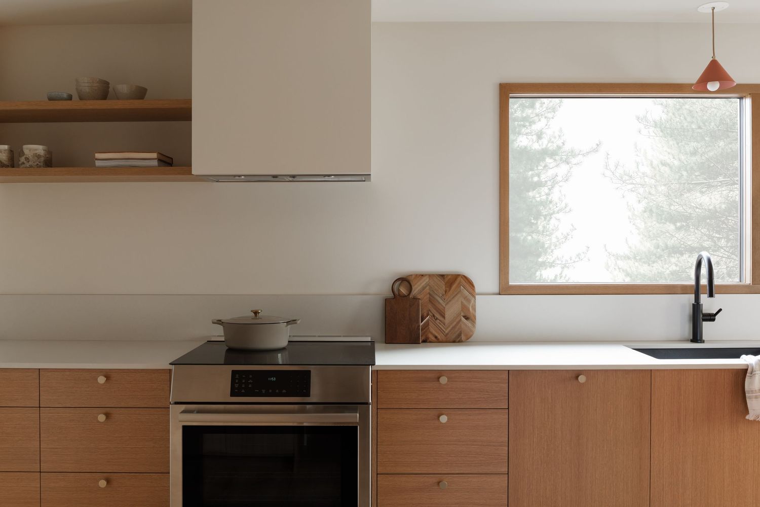Minimalist wood kitchen with white countertops, black faucet, induction range, and framed window overlooking snowy trees.
