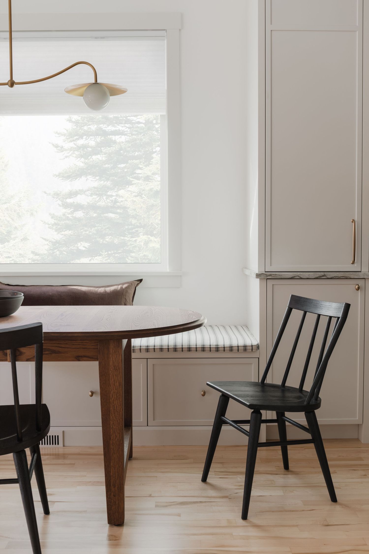 Minimalist dining nook with dark wood round table, black spindle chairs, built-in bench, and brass pendant light.