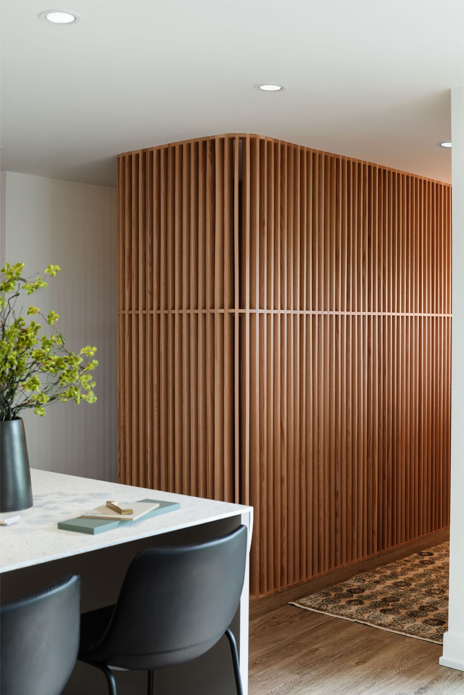 Contemporary kitchen with white marble island, black chairs, and curved warm wood slatted accent wall divider.