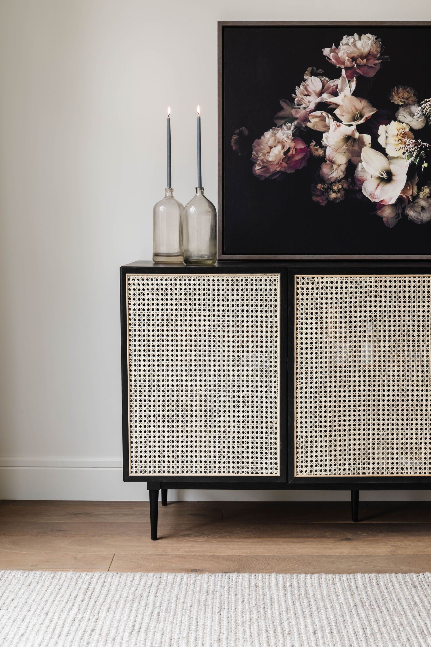Black cane sideboard with glass candleholders and dark floral artwork on light wall.