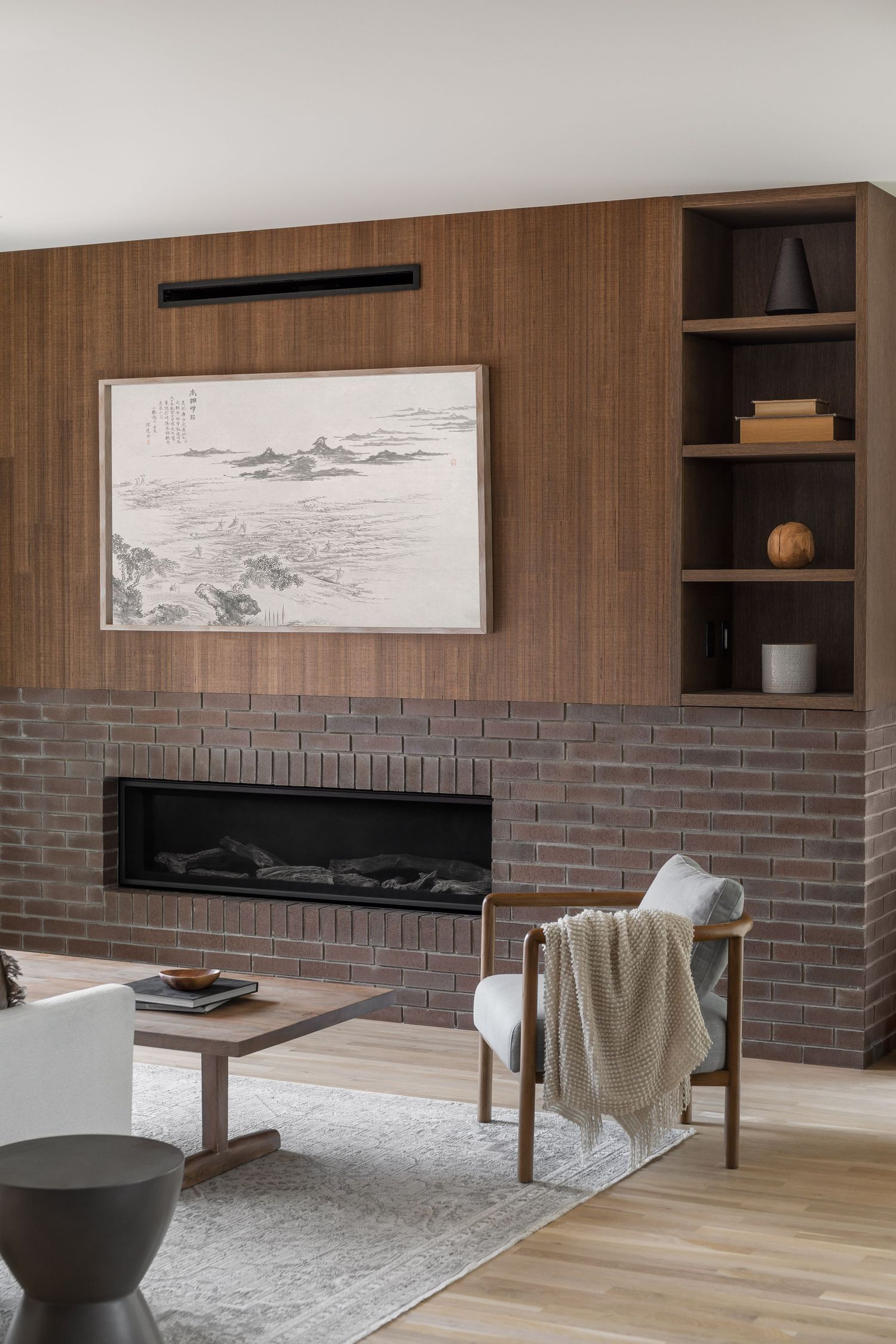 Modern living room with dark brick fireplace, wood panel wall, built-in shelving, and Asian landscape artwork.