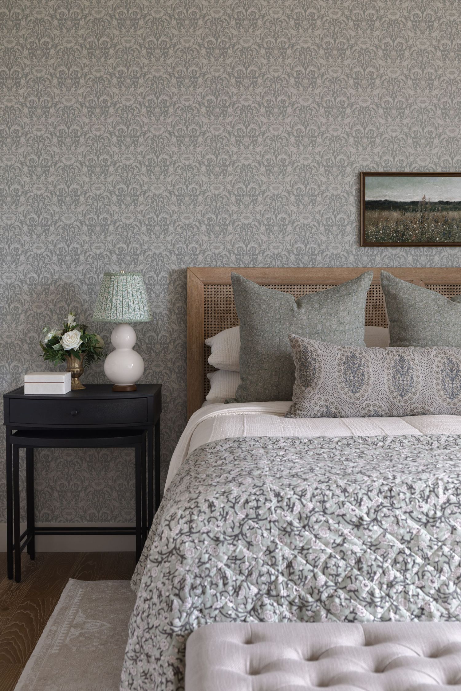 Elegant bedroom with gray floral wallpaper, cane headboard, layered patterned bedding, and black nightstand.