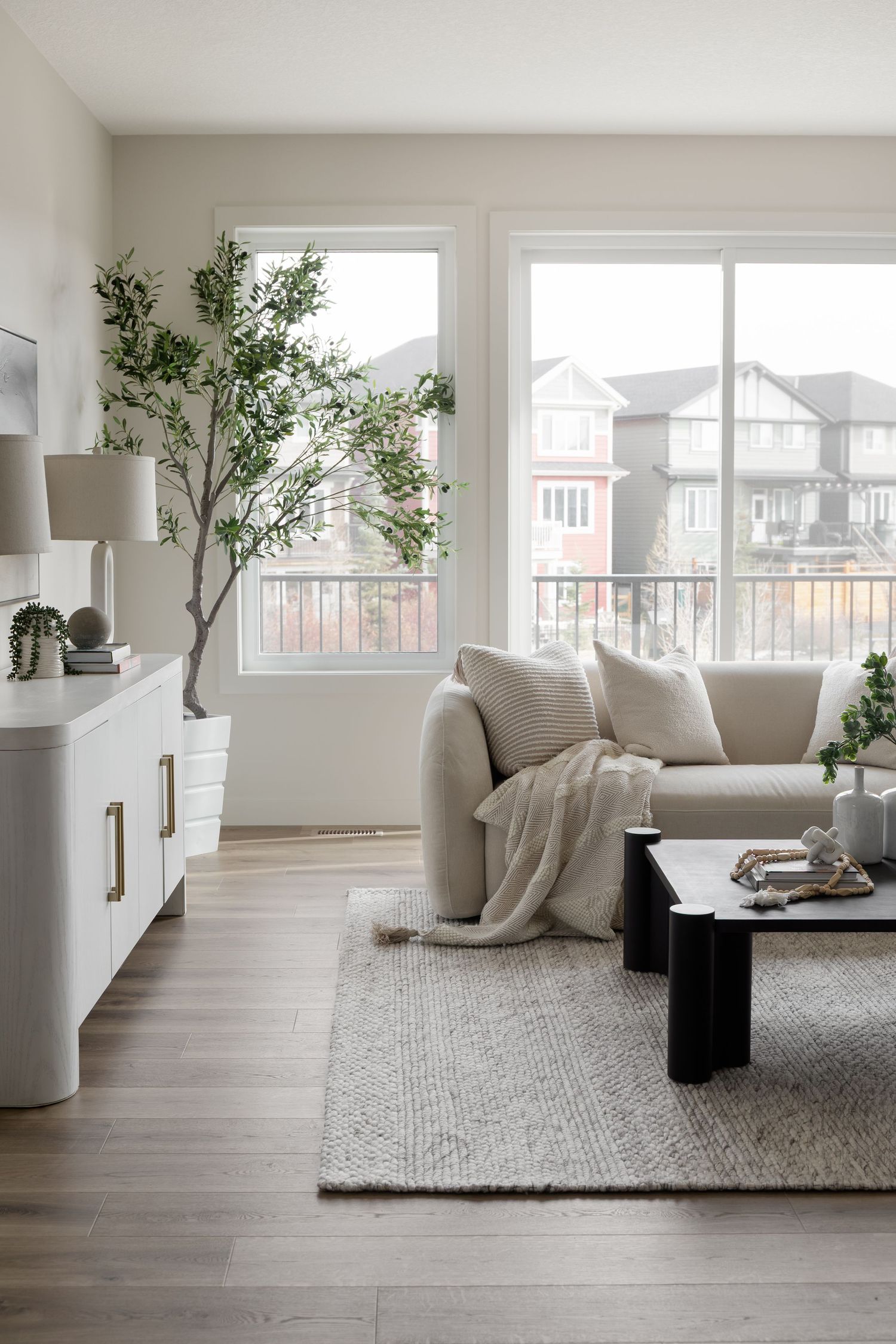 Bright minimalist living room with beige sofa, gray rug, black coffee table, white sideboard, and indoor tree by large windows.