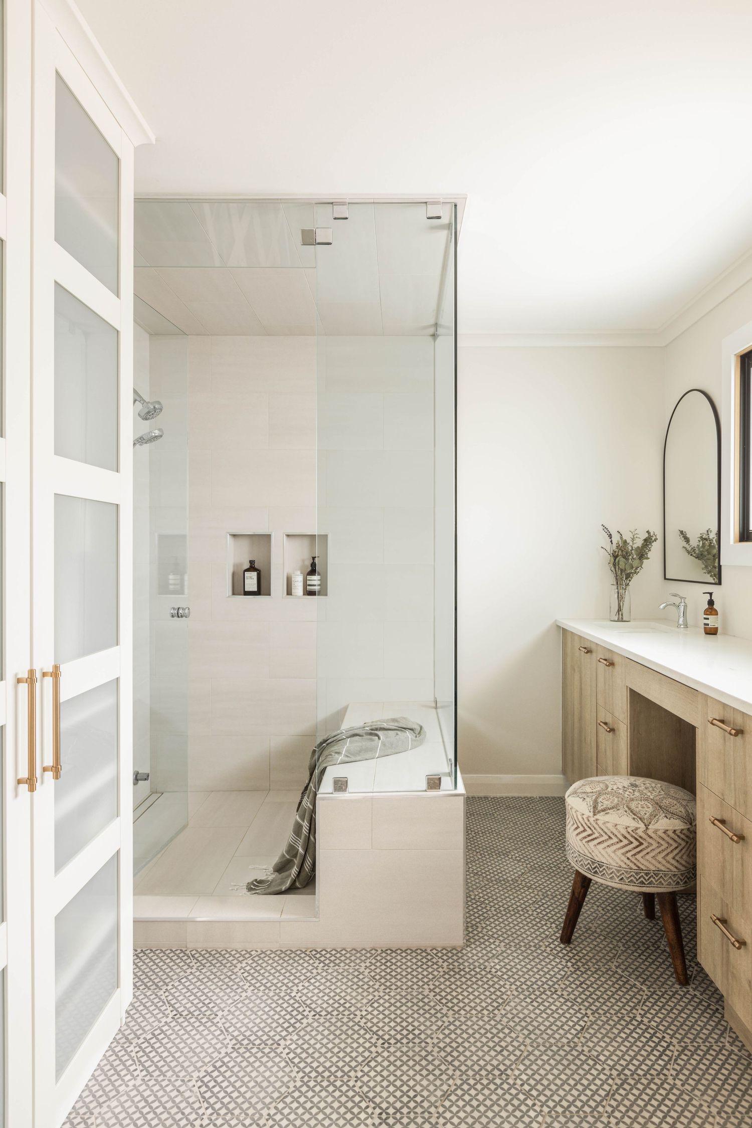 Bright bathroom with glass enclosed shower, penny tile floor, light wood vanity, arched mirror, and French doors.