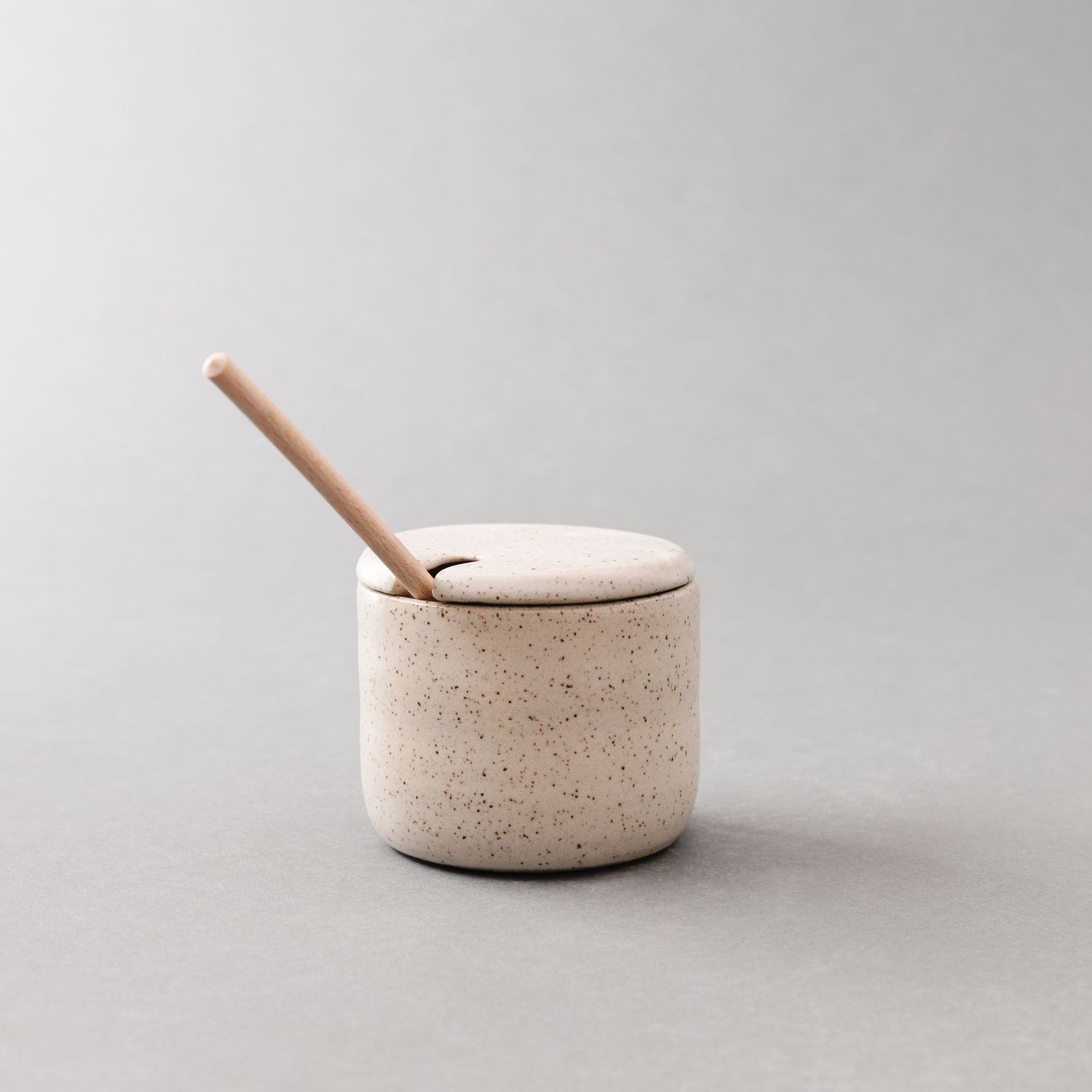 Speckled ceramic sugar pot with wooden spoon resting on lid, minimalist Japandi style stoneware in neutral beige tones.
