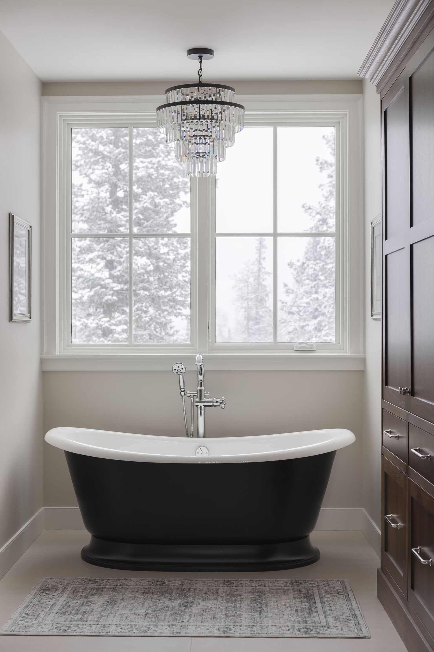 Luxurious black freestanding soaking tub beneath crystal chandelier with snowy window view and dark wood cabinetry.
