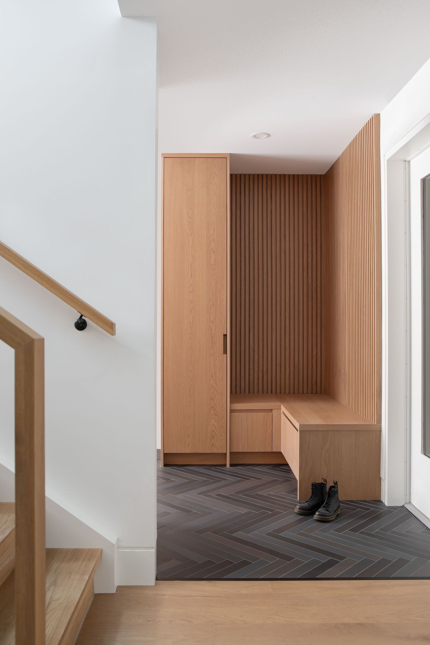 Modern entryway with natural oak wood built-in cabinetry, slatted wall panels, bench seating, and herringbone dark tile flooring.