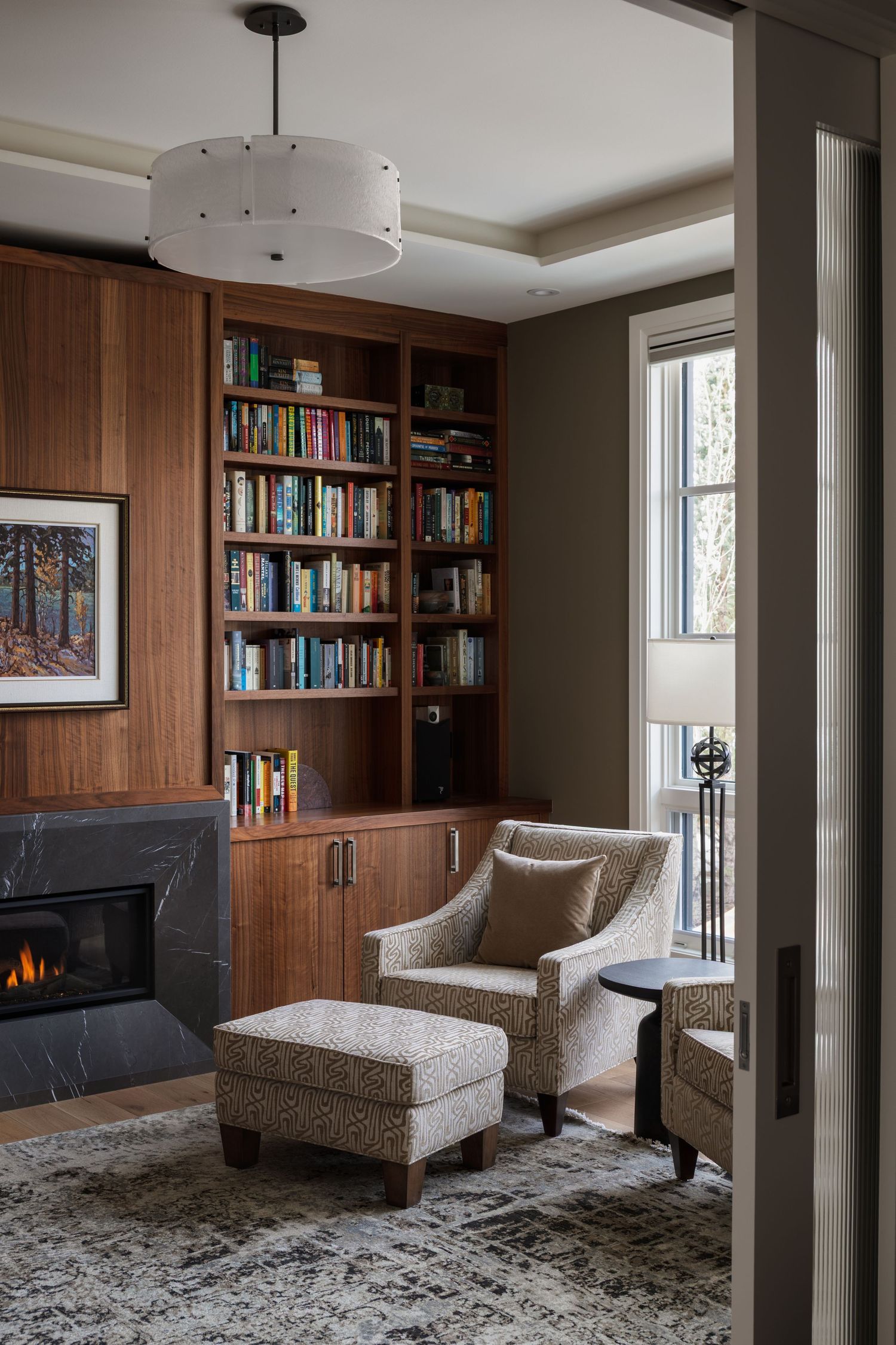 Cozy reading nook with walnut built-in bookshelves, marble fireplace, patterned armchair with ottoman, and drum pendant light.