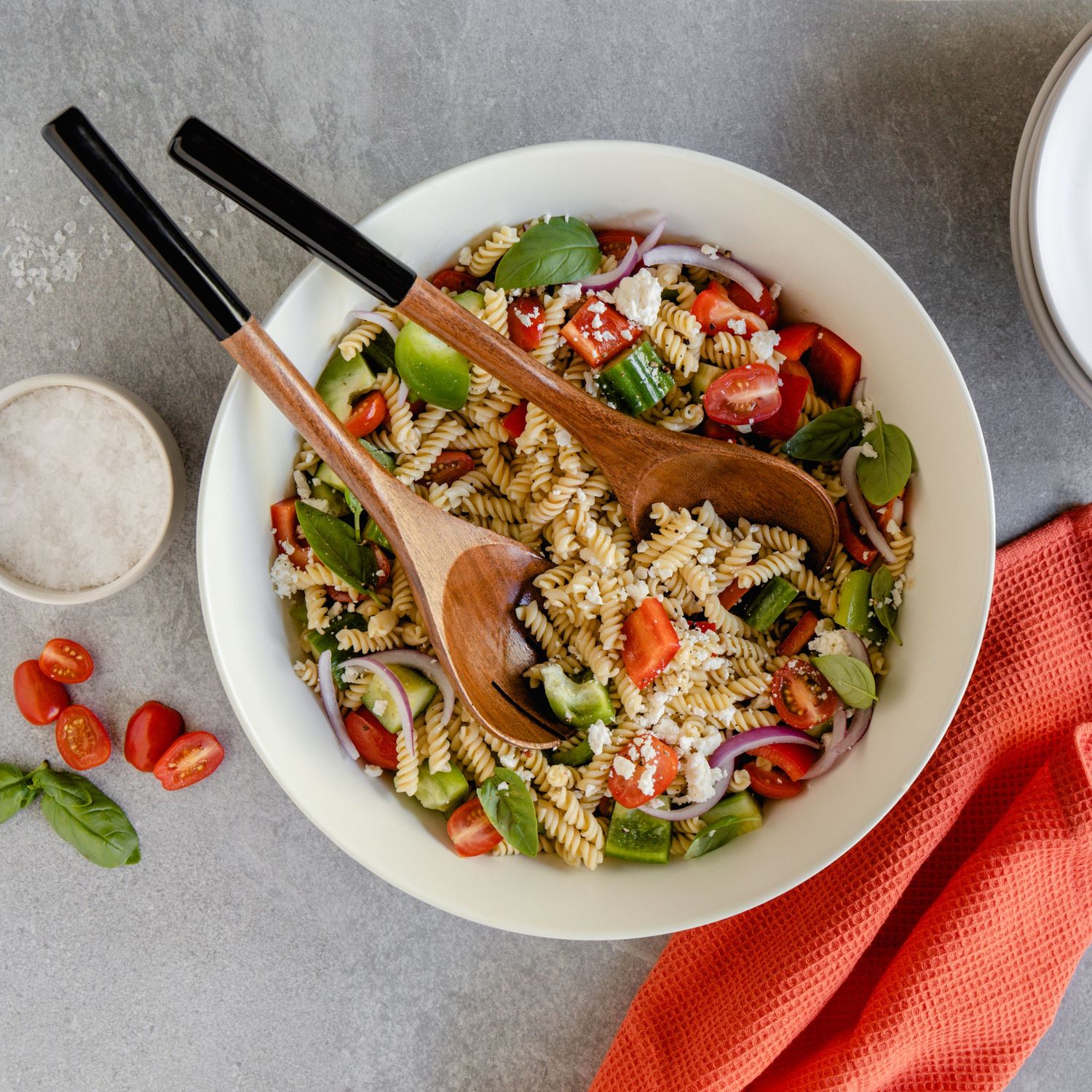 Colorful rotini pasta salad with cherry tomatoes, spinach, and red onion served in a white bowl with wooden spoons.
