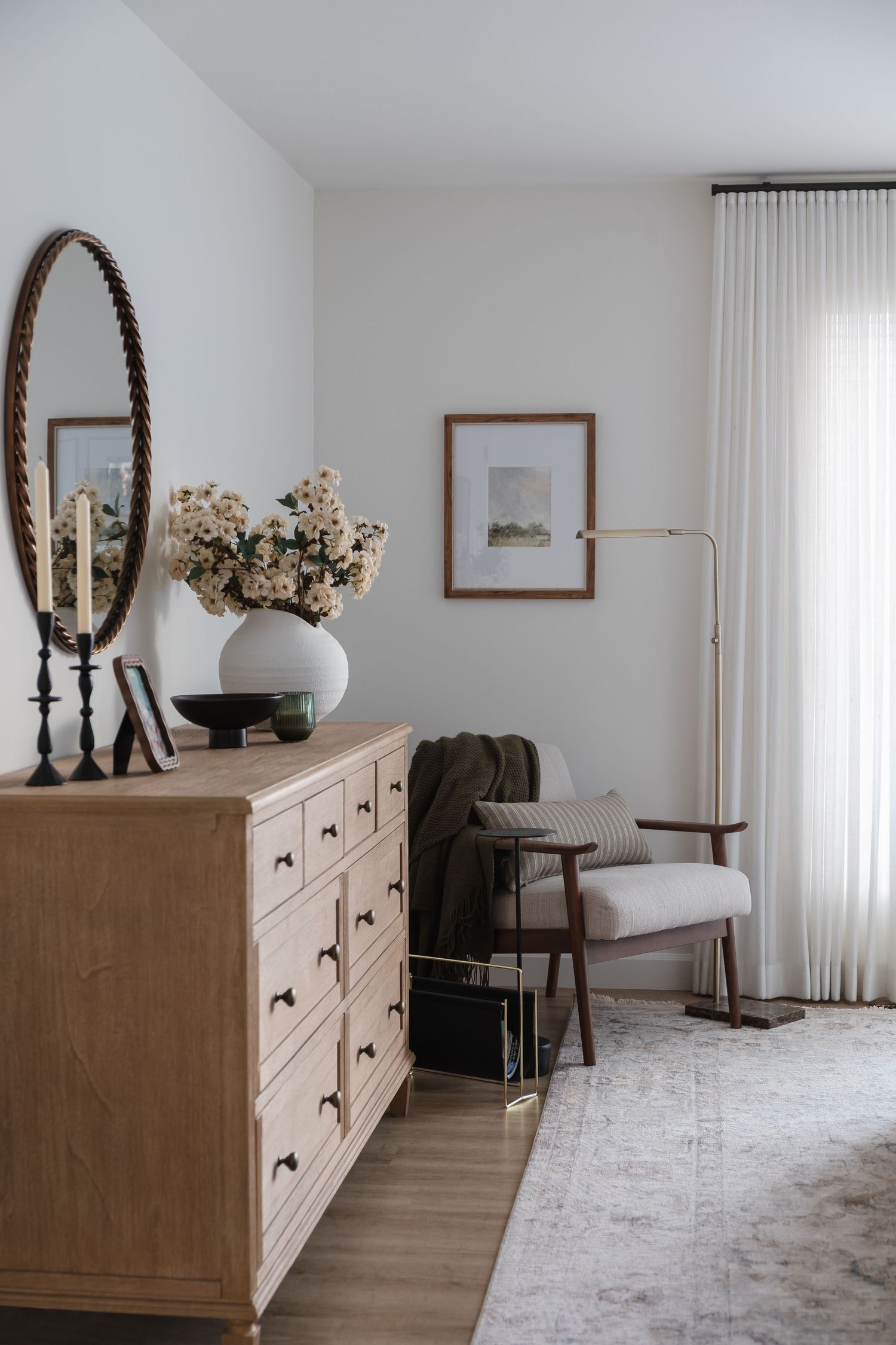Bright bedroom corner with oak dresser, oval mirror, white floral vase, and neutral accent chair.