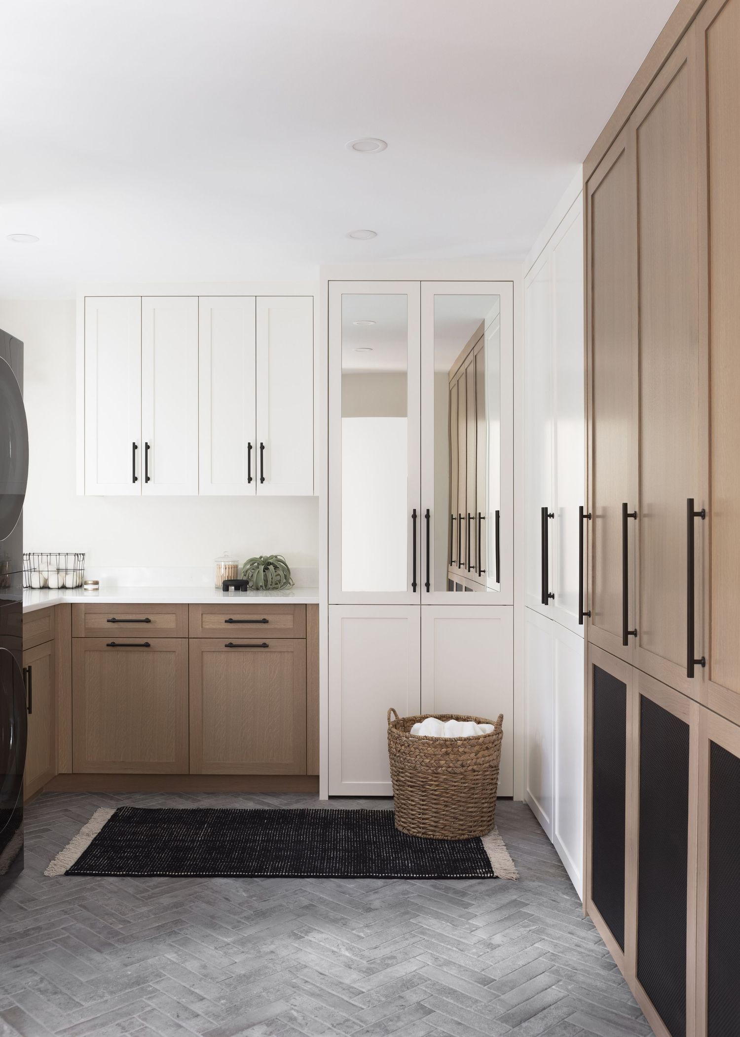 Modern mudroom with two-tone white and light wood cabinetry, mirrored cabinet, black hardware, and gray herringbone tile floor.