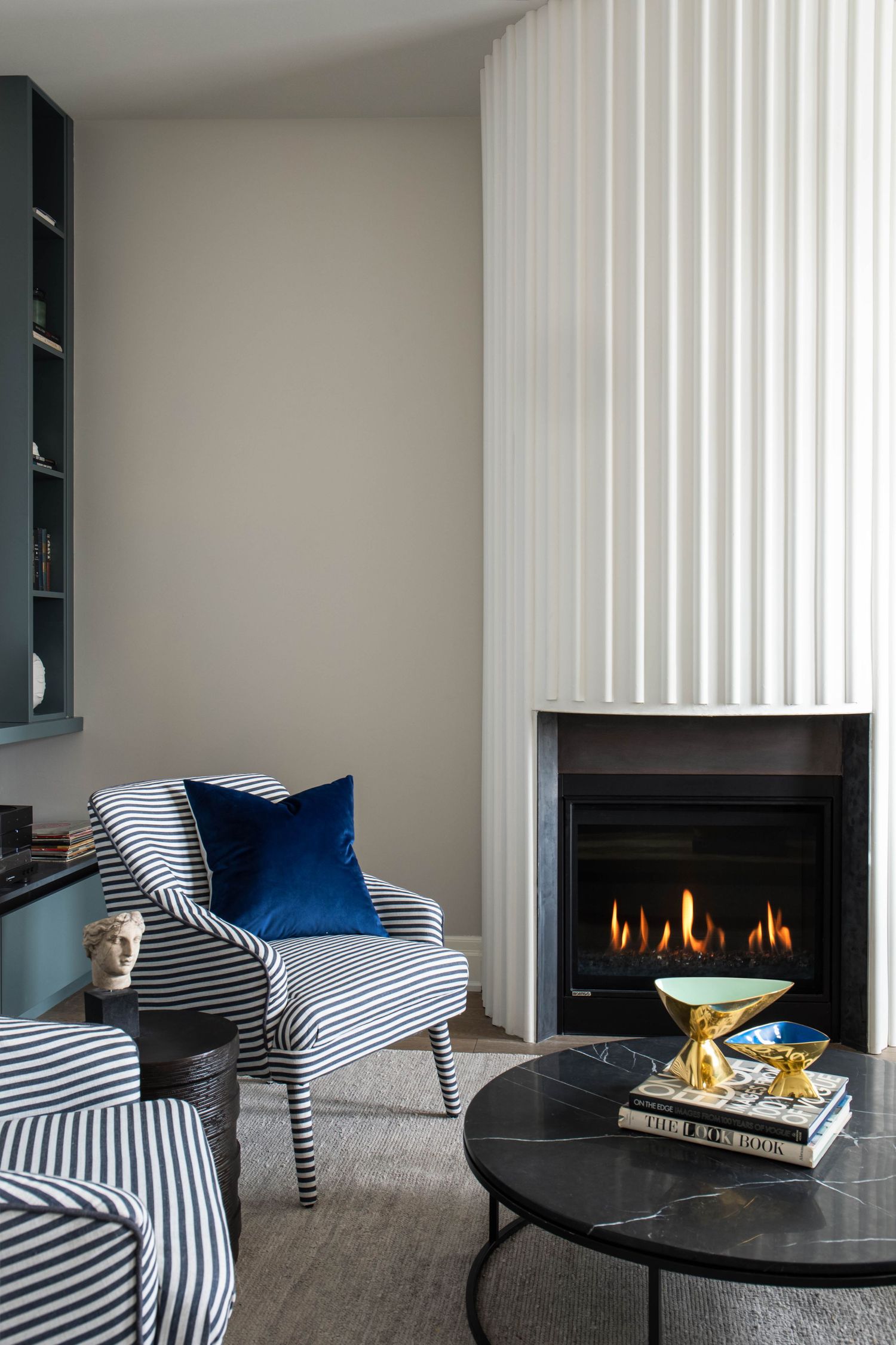 Cozy living room corner with striped armchair, blue velvet pillow, marble coffee table, and fluted white fireplace.
