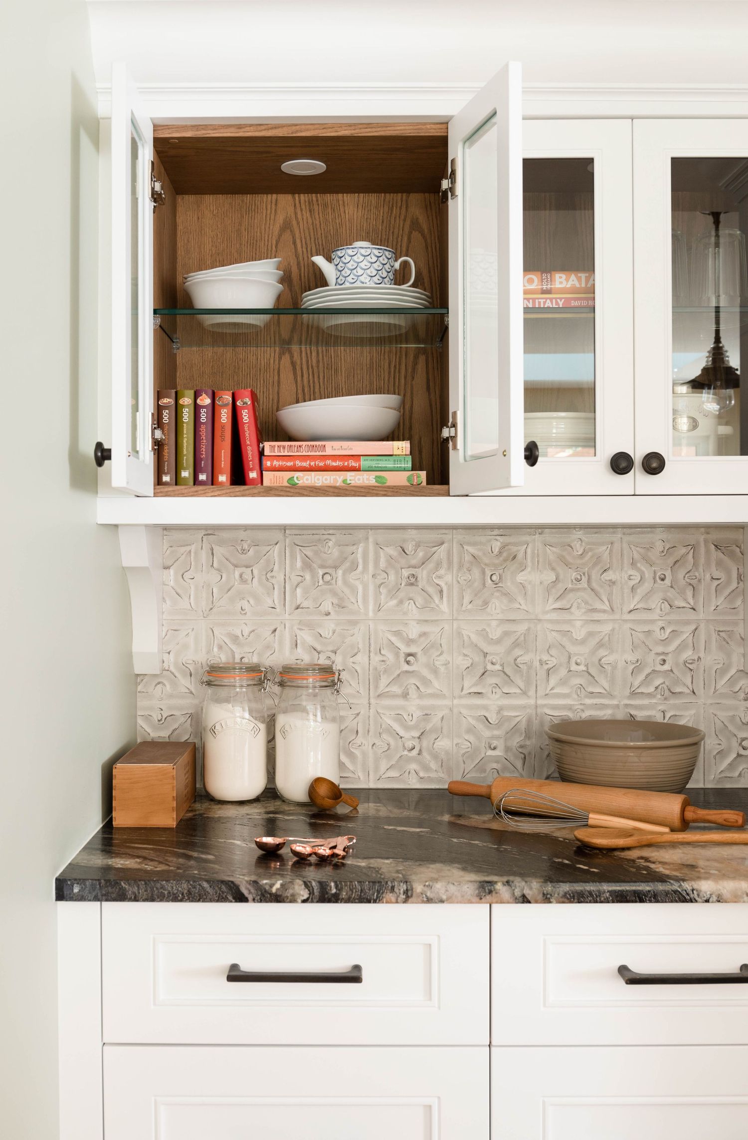 White kitchen with ornate tile backsplash, dark granite counters, open cabinet displaying dishes and cookbooks.