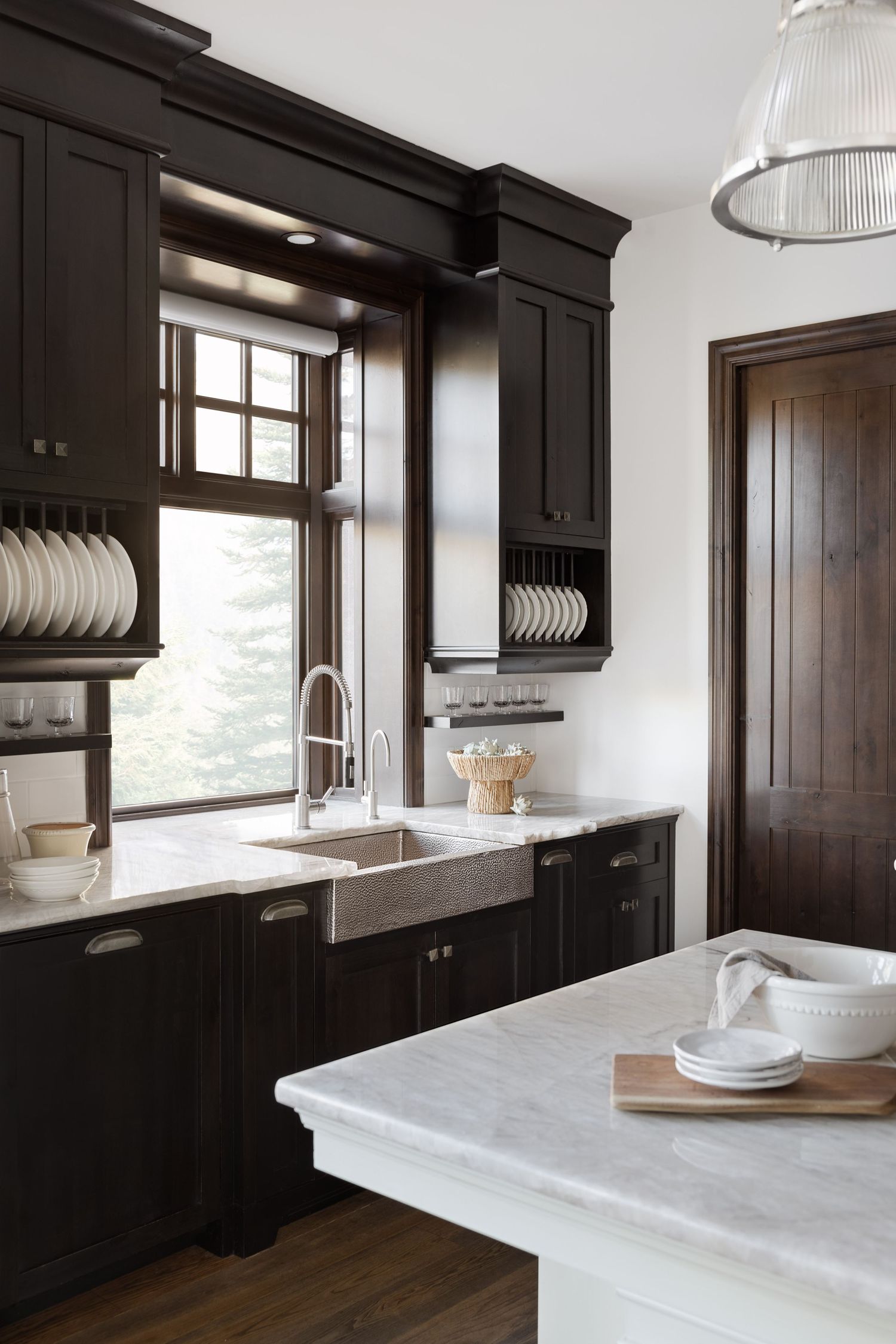 Dramatic dark kitchen with black cabinets, marble island, farmhouse sink, plate rack shelves, and pendant light.