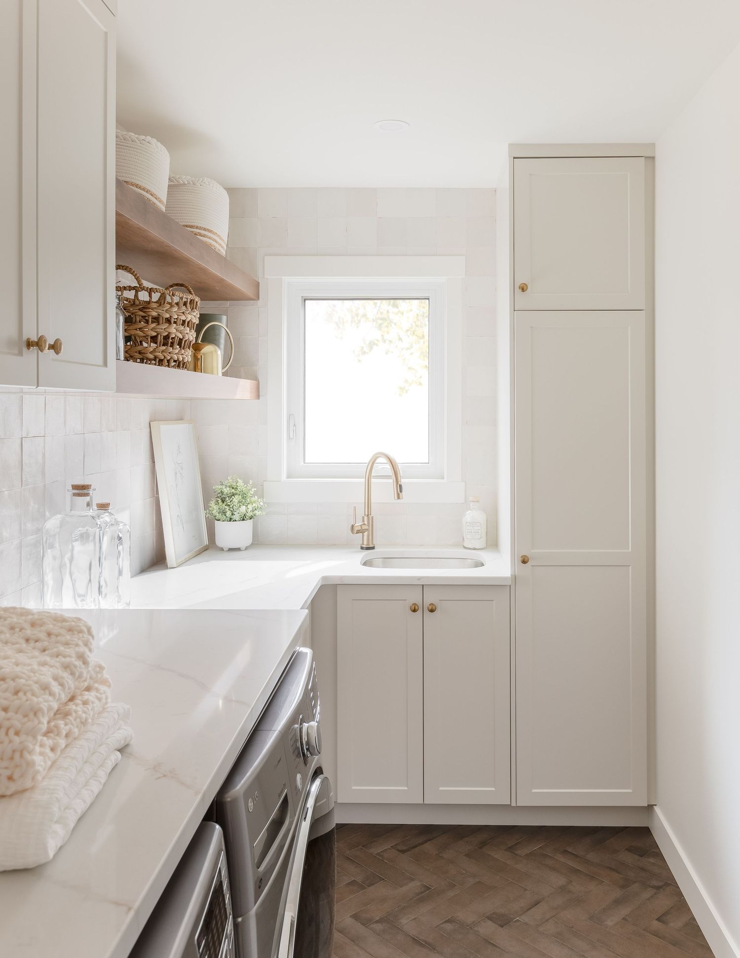 Bright laundry room with greige shaker cabinets, herringbone floors, and rose gold faucet.