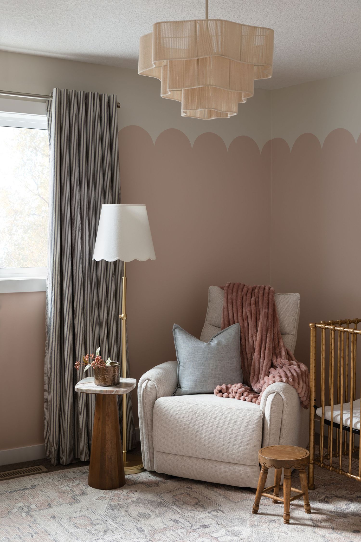 Cozy nursery corner with blush scallop walls, cream glider, gold crib, layered throw pillows, floor lamp, and tiered chandelier.