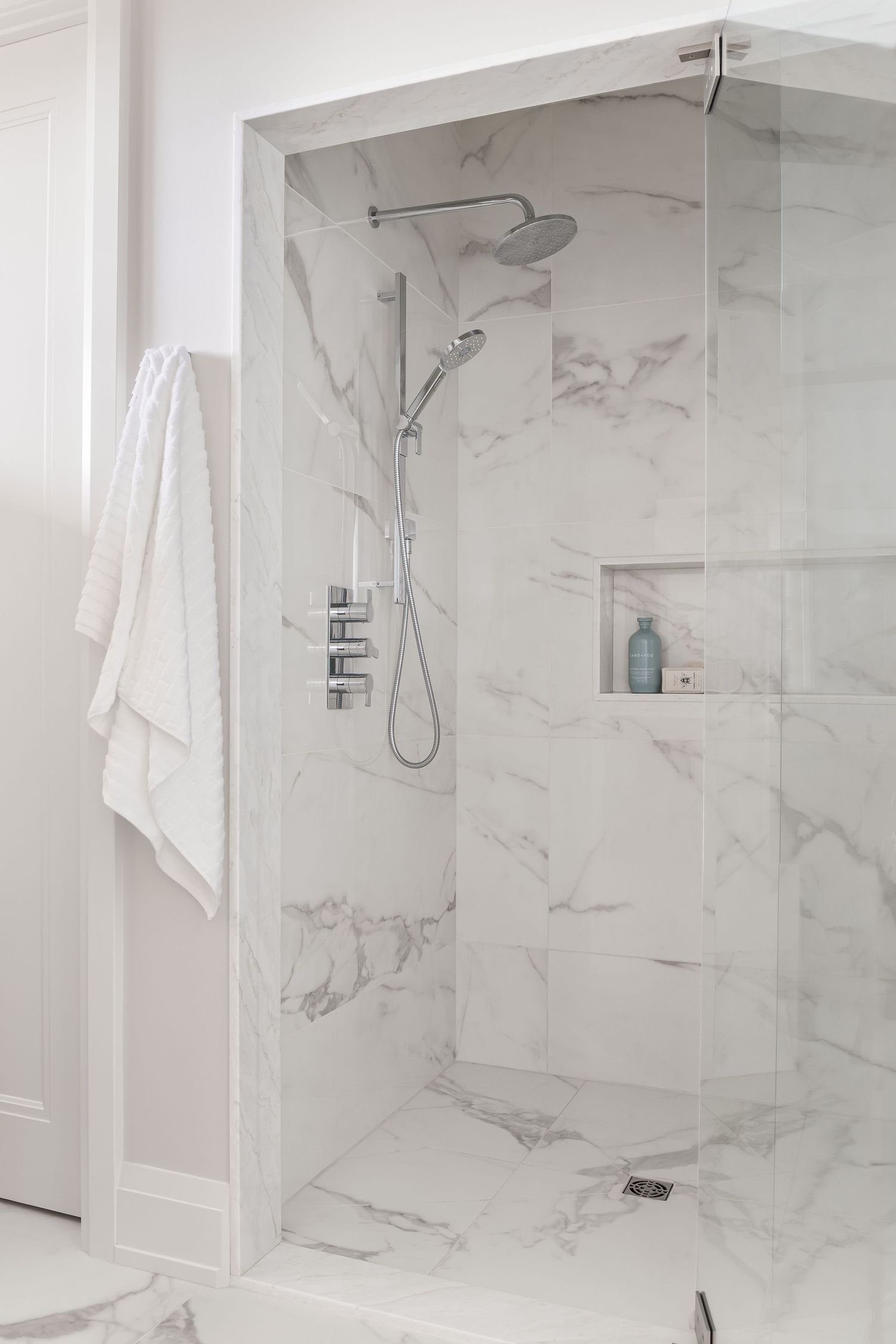 Luxurious walk-in shower with white marble tiles, chrome fixtures, rainfall showerhead, and built-in niche shelf.