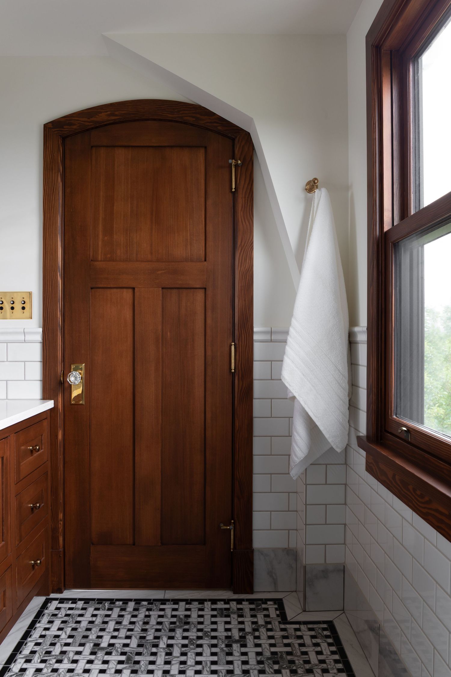 Craftsman bathroom with arched walnut door, brass hardware, subway tile walls, mosaic floor, and white towel on hook.