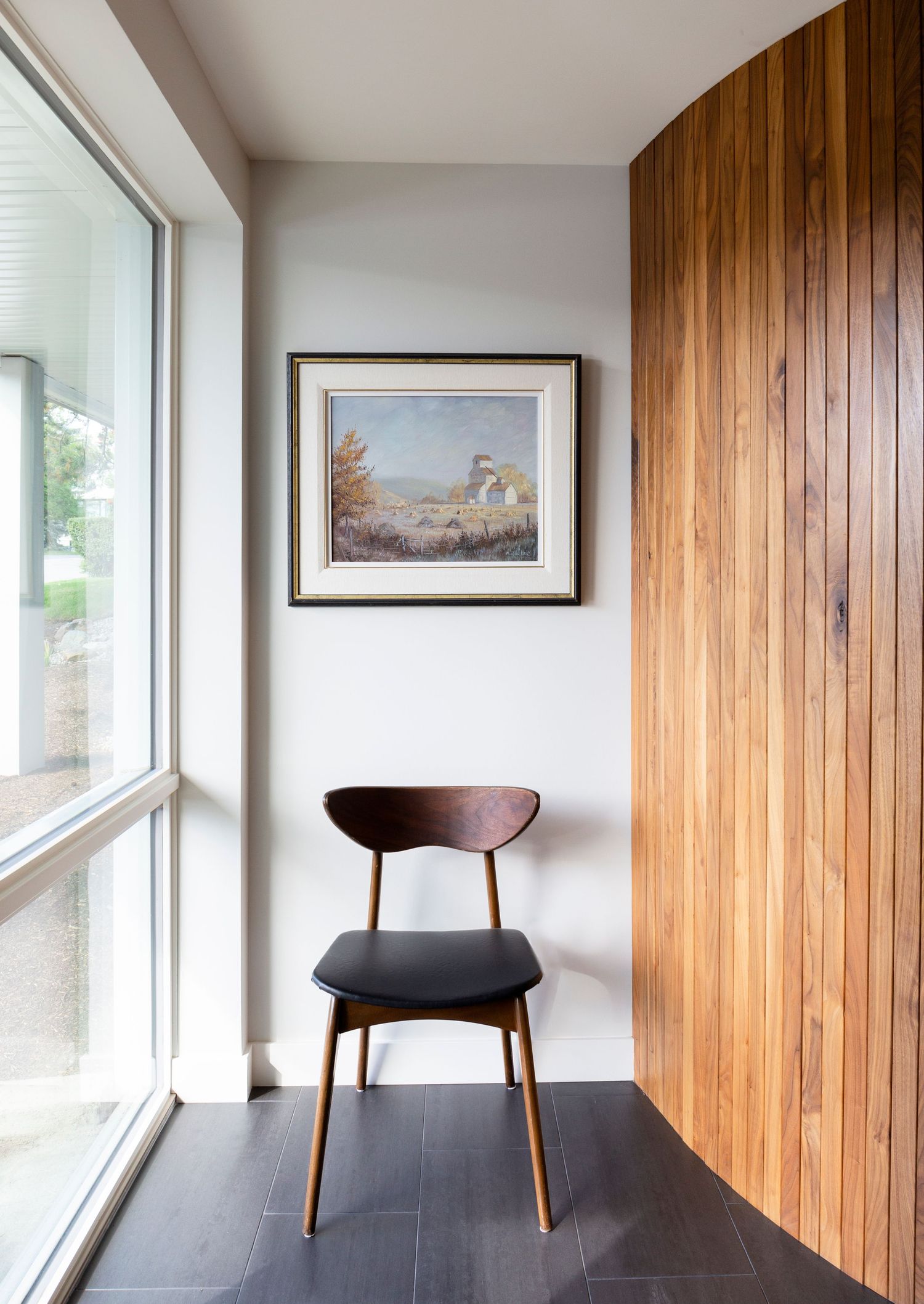 Mid-century modern wooden chair beside large window with curved wood panel wall accent.