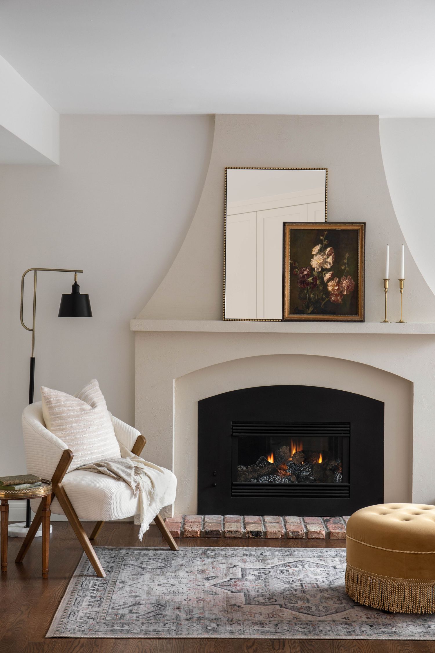 Cozy living room corner with arched white fireplace, bouclé chair, mustard ottoman, and framed artwork on mantel.