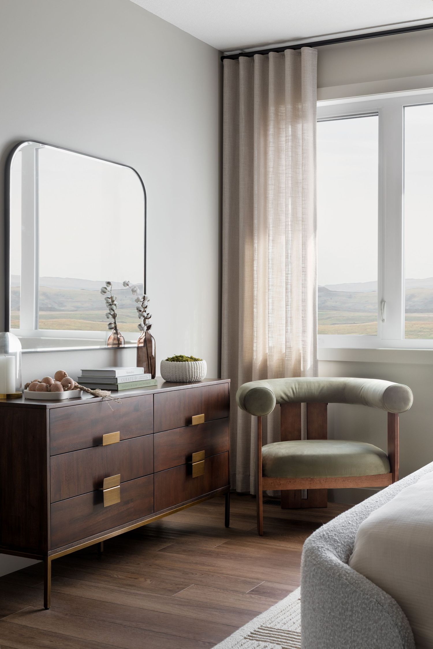 Contemporary bedroom corner with dark wood dresser, arched mirror, velvet accent chair, and scenic window view.